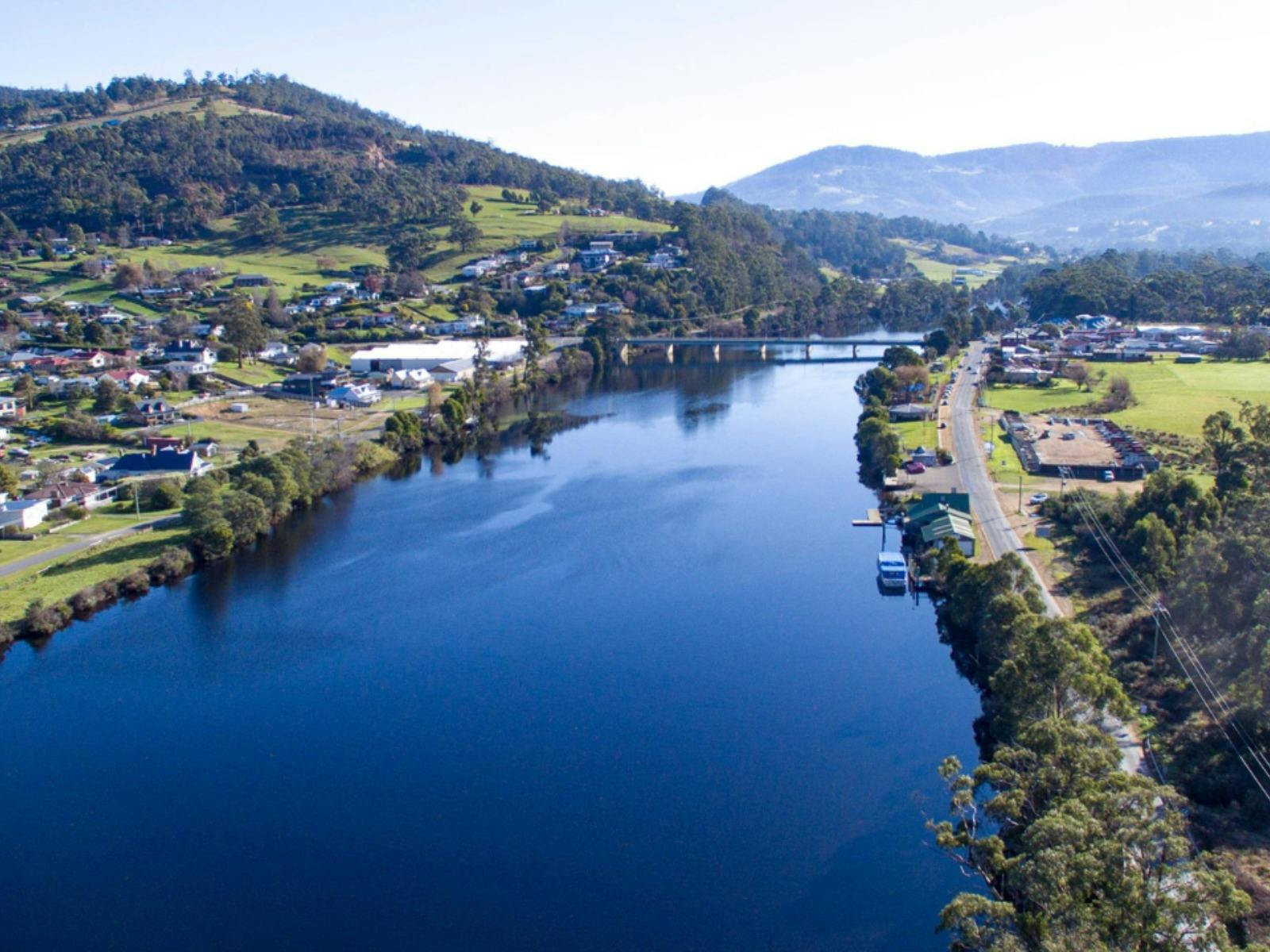 Drone shot of a wide, dark blue river flowing through a green, wooded town.