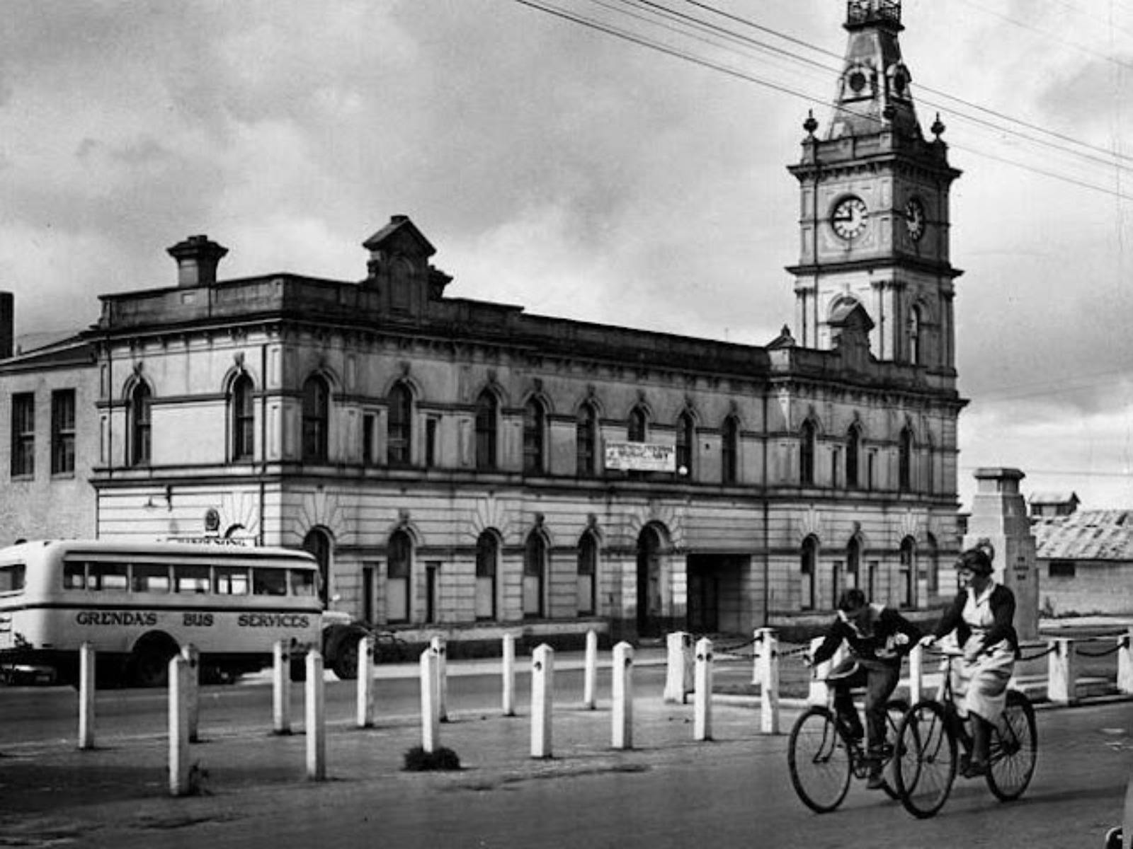 Black and white photo of historical Drum Theatre building with two cyclists riding by.