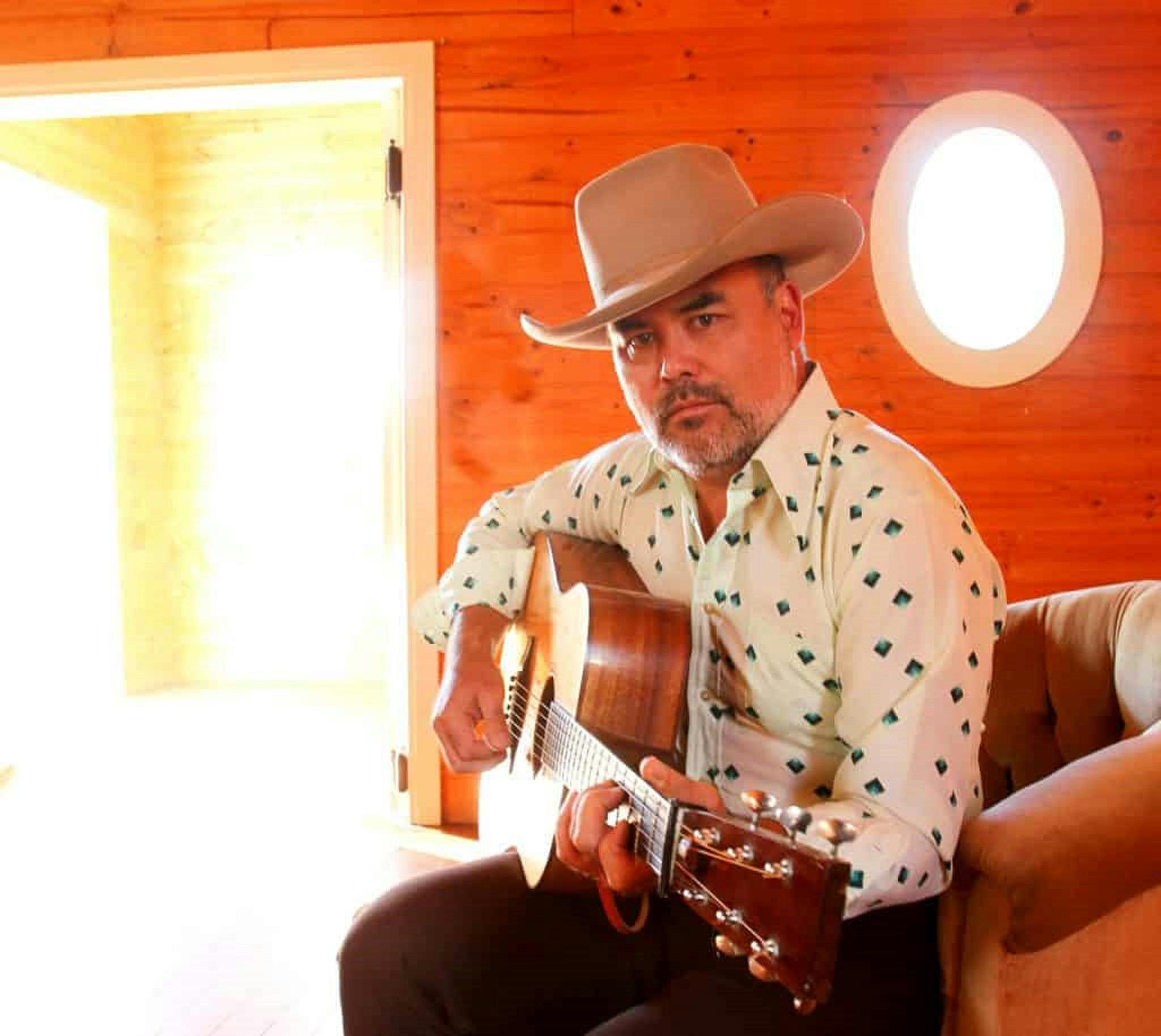 Pete Denahy on a couch, wearing a cowboy hat, strumming a guitar looking directly at the camera