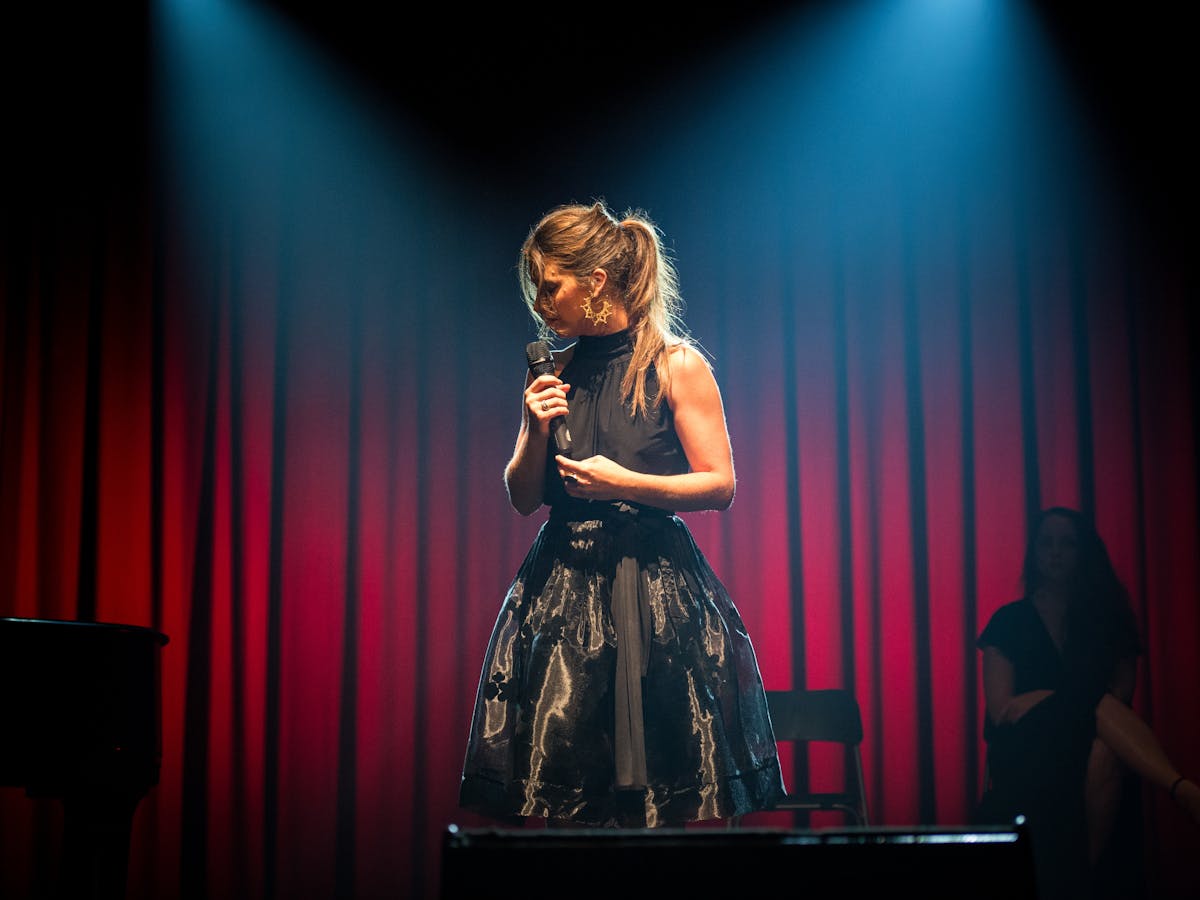 Female in black dress on stage with head to the side, holding microphone, in front of red curtians.