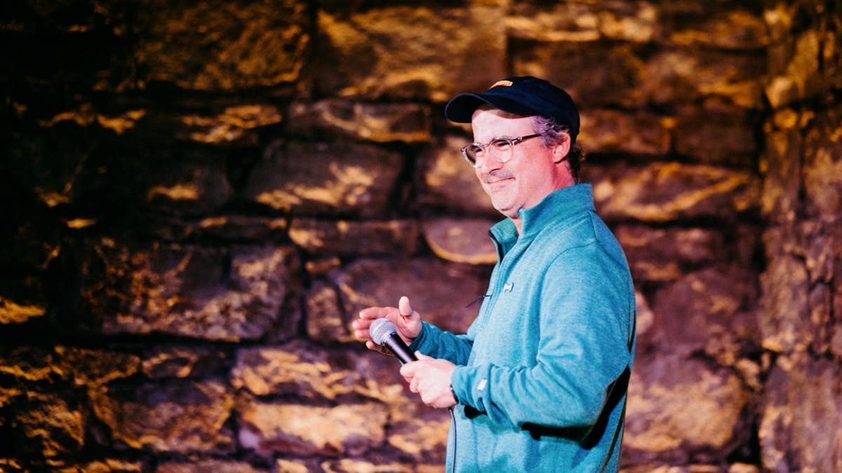 Comedian Stu Daulman on stage at a comedy club, with a dimly backlit heritage brick wall behind him Comedian Stu Daulman on stage at a comedy club, with a dimly backlit heritage brick wall behind him