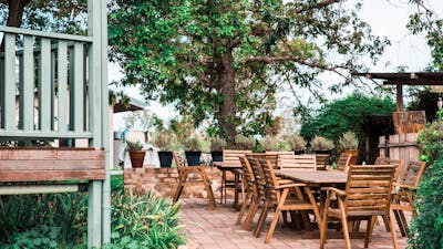 Timber tables and chairs arranged on a paved area underneath a large tree.