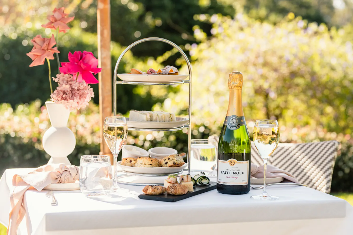 A high tea stand filled with treats and a bottle of champagne on a white table in the garden