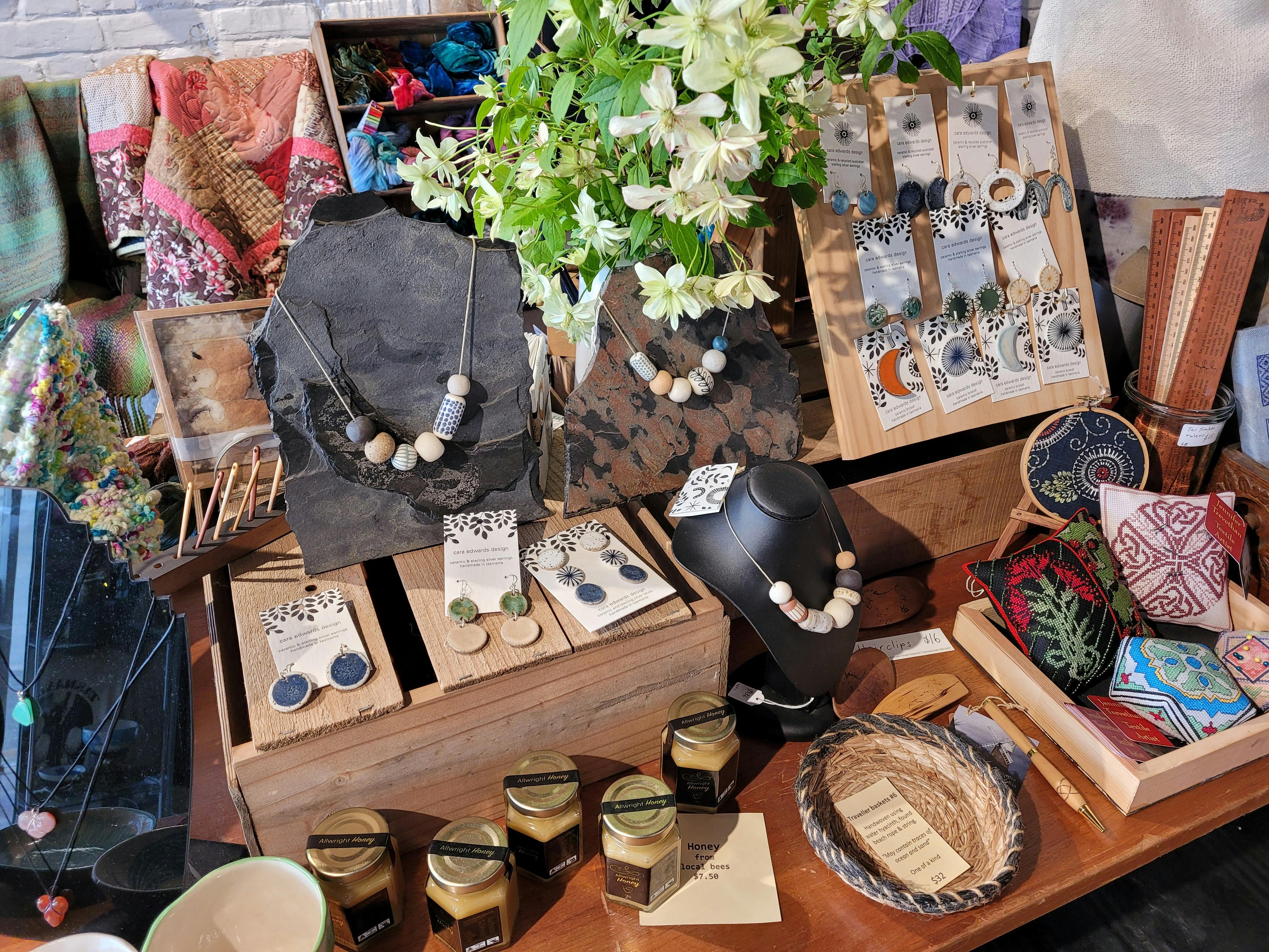 A display of ceramic jewellery, woven baskets, jars of honey and stitched pin cushions on a table.
