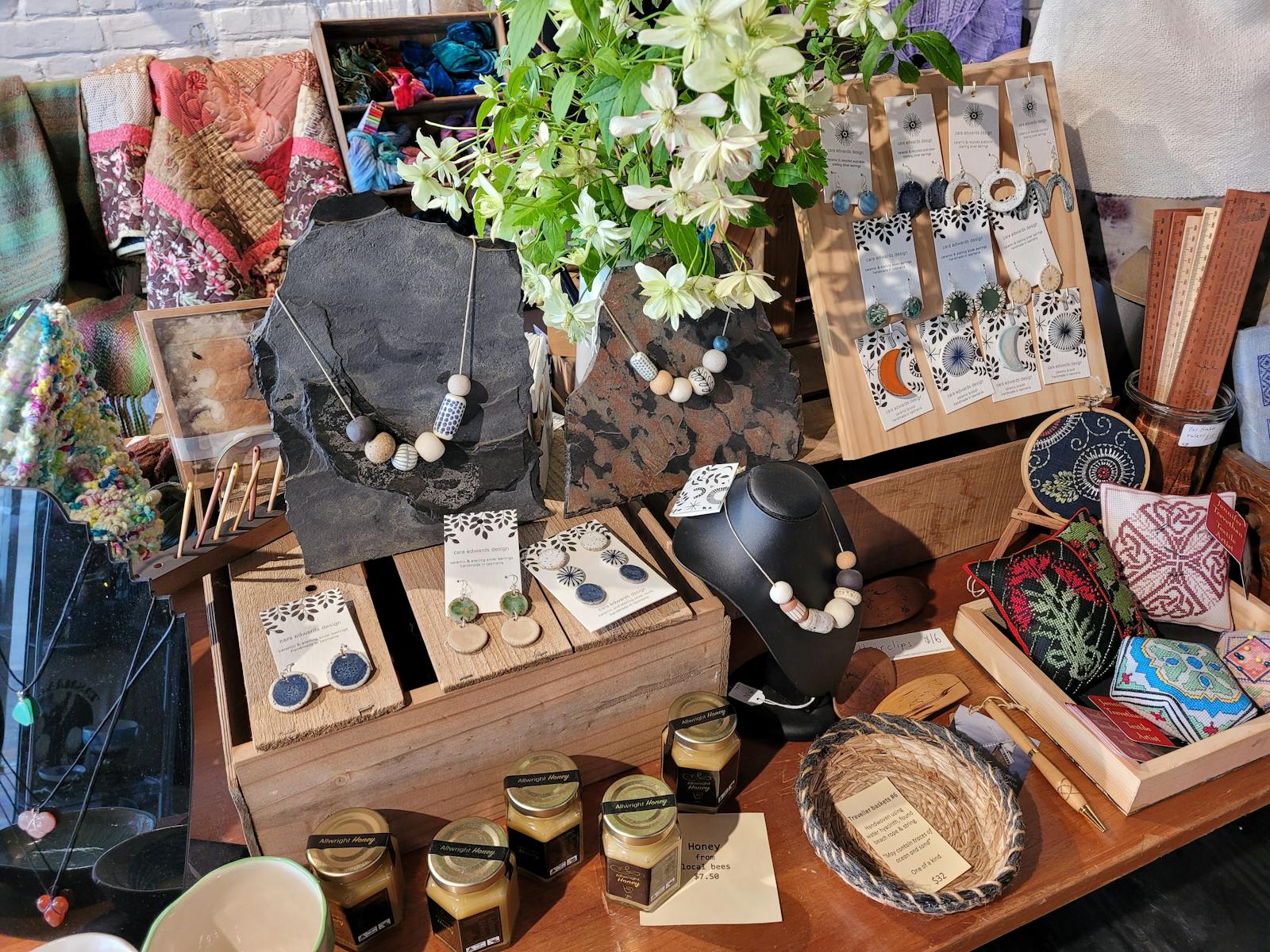 A display of ceramic jewellery, woven baskets, jars of honey and stitched pin cushions on a table.