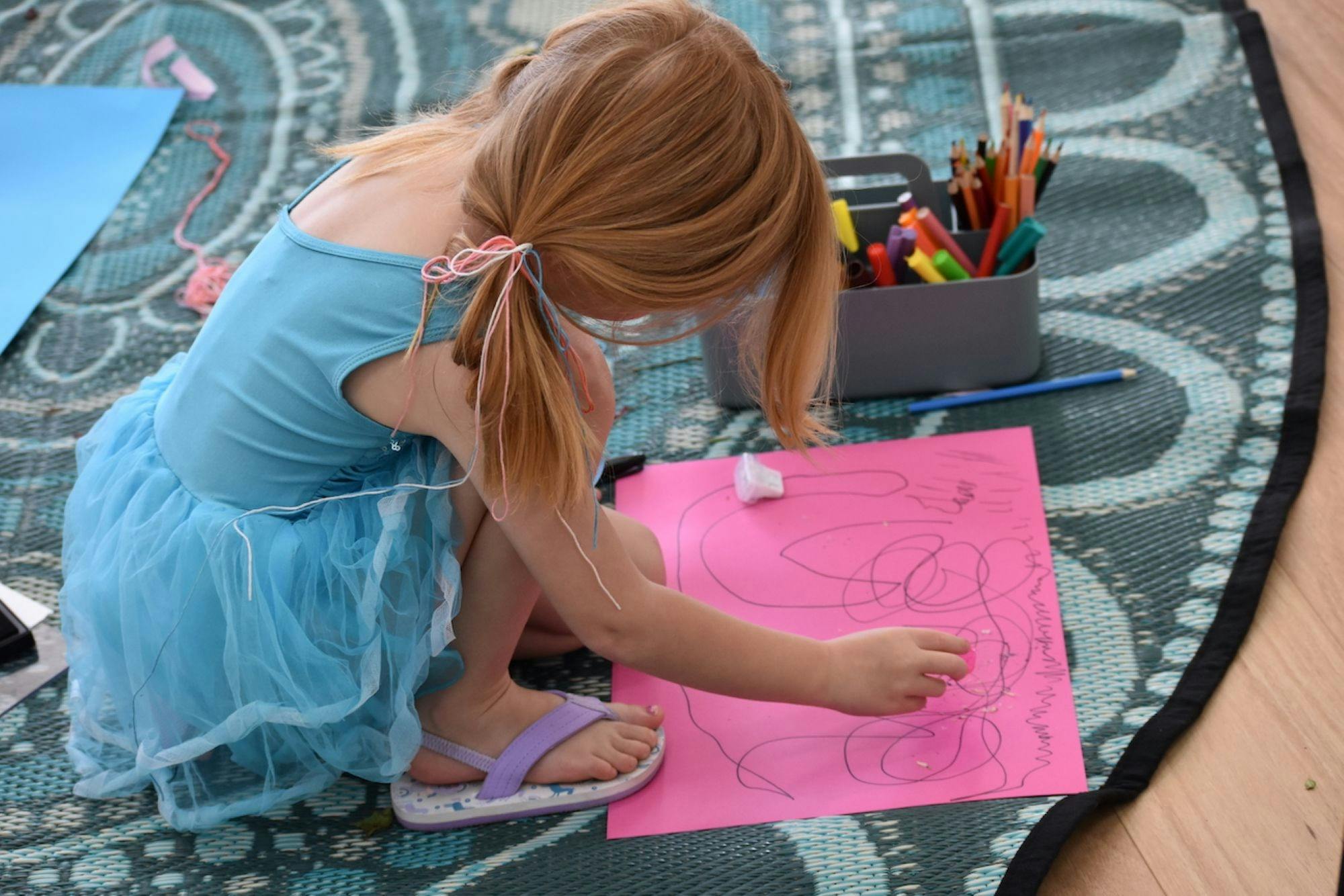 Small girl in blue dress sitting on a rug drawing pictures on a piece of bright pink paper