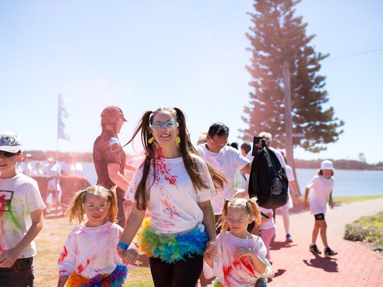 Lady stands between 2 small children, holding hands, and are covered in rainbow coloured chalk