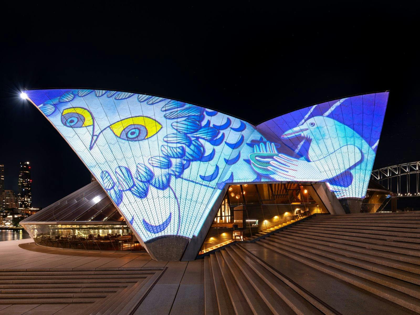 The Opera House western sails lit with  a blue and white owl with yellow eyes and a white raven