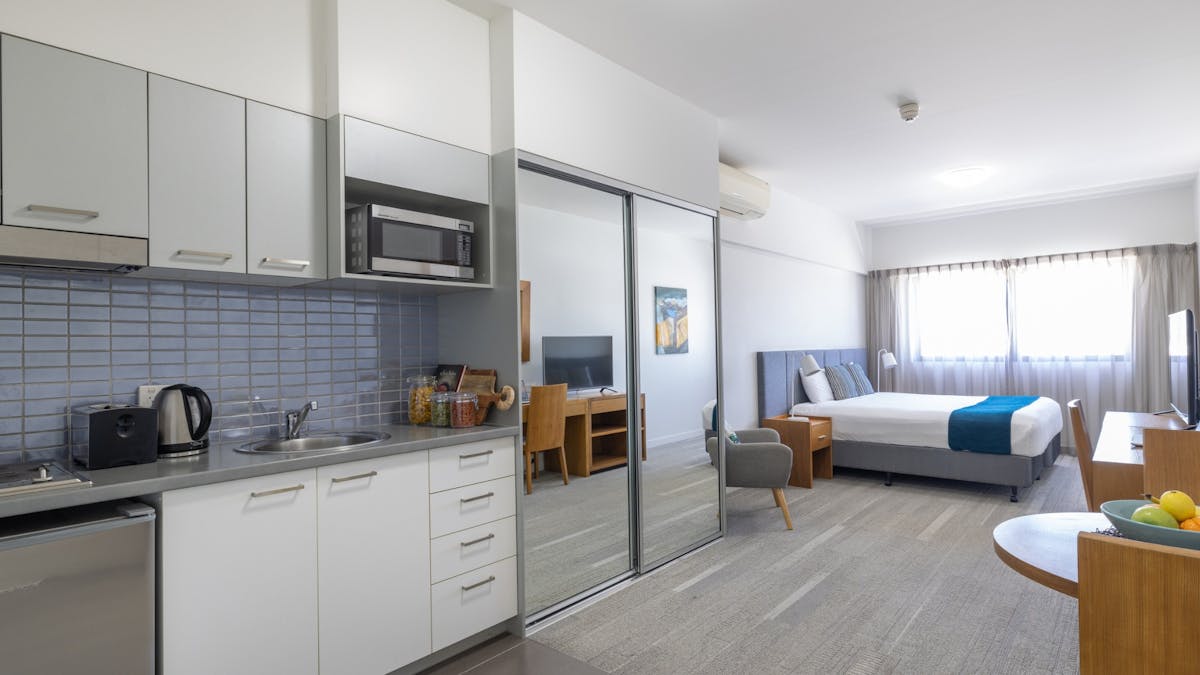 Interior of a studio apartment with kitchen cupboards, bench, large mirrors and bed.