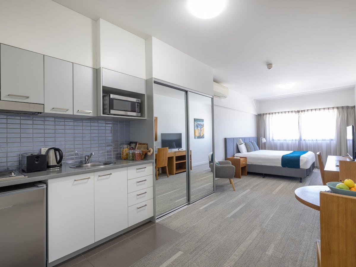 Interior of a studio apartment with kitchen cupboards, bench, large mirrors and bed.