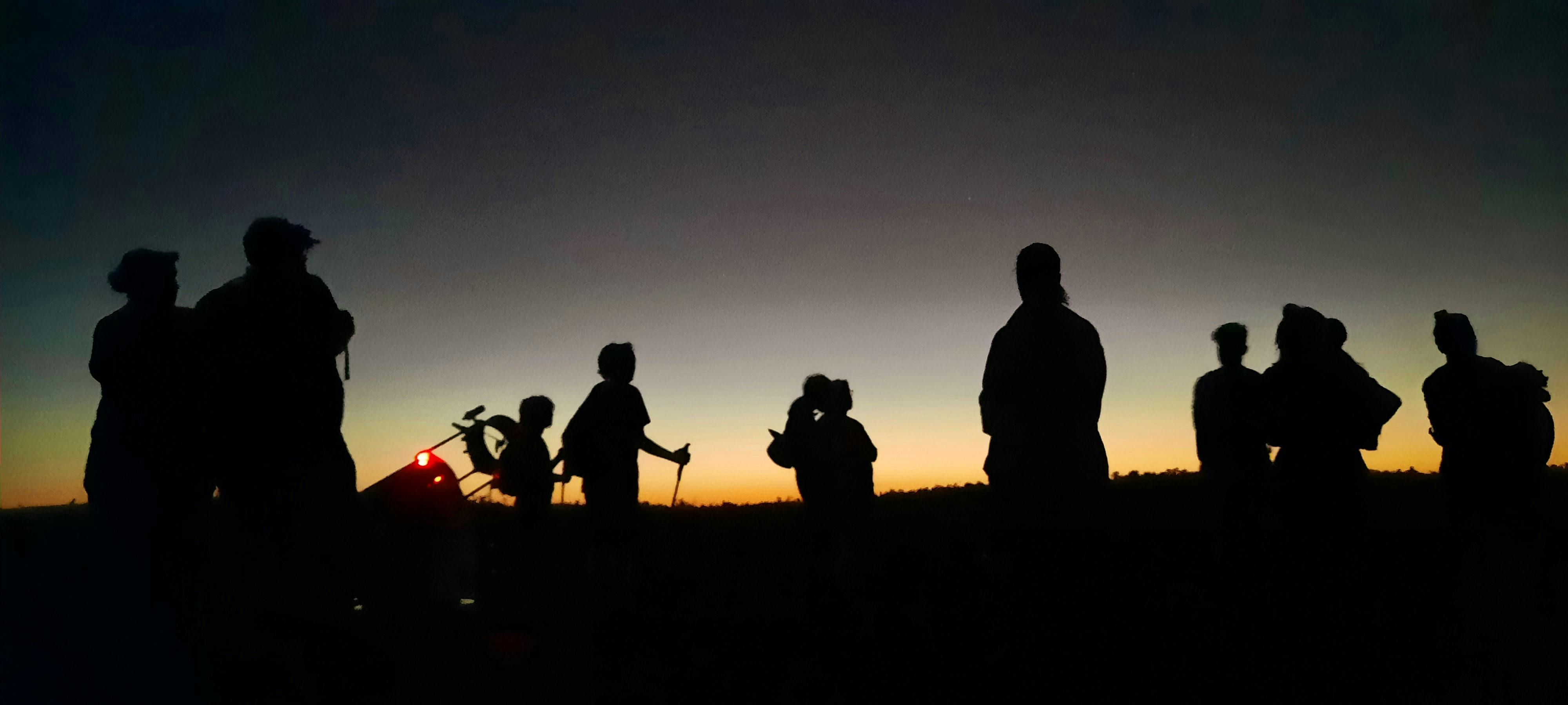 Silhouettes of people looking up at the stars at dusk, deep purple hues in sky warm tones on horizon