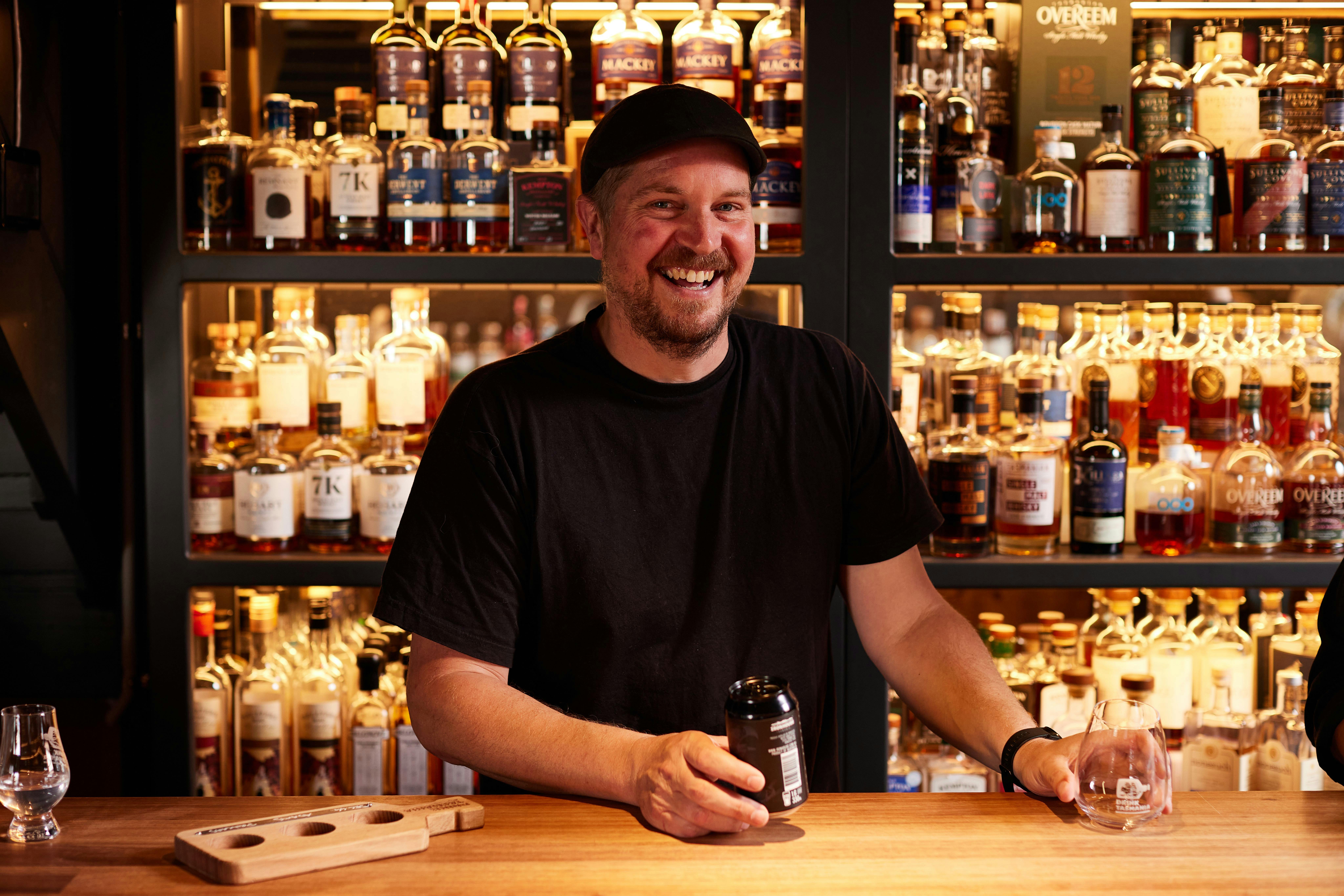 Smiling staff member about to pour a drink with shelves of delicious spirits behind them.
