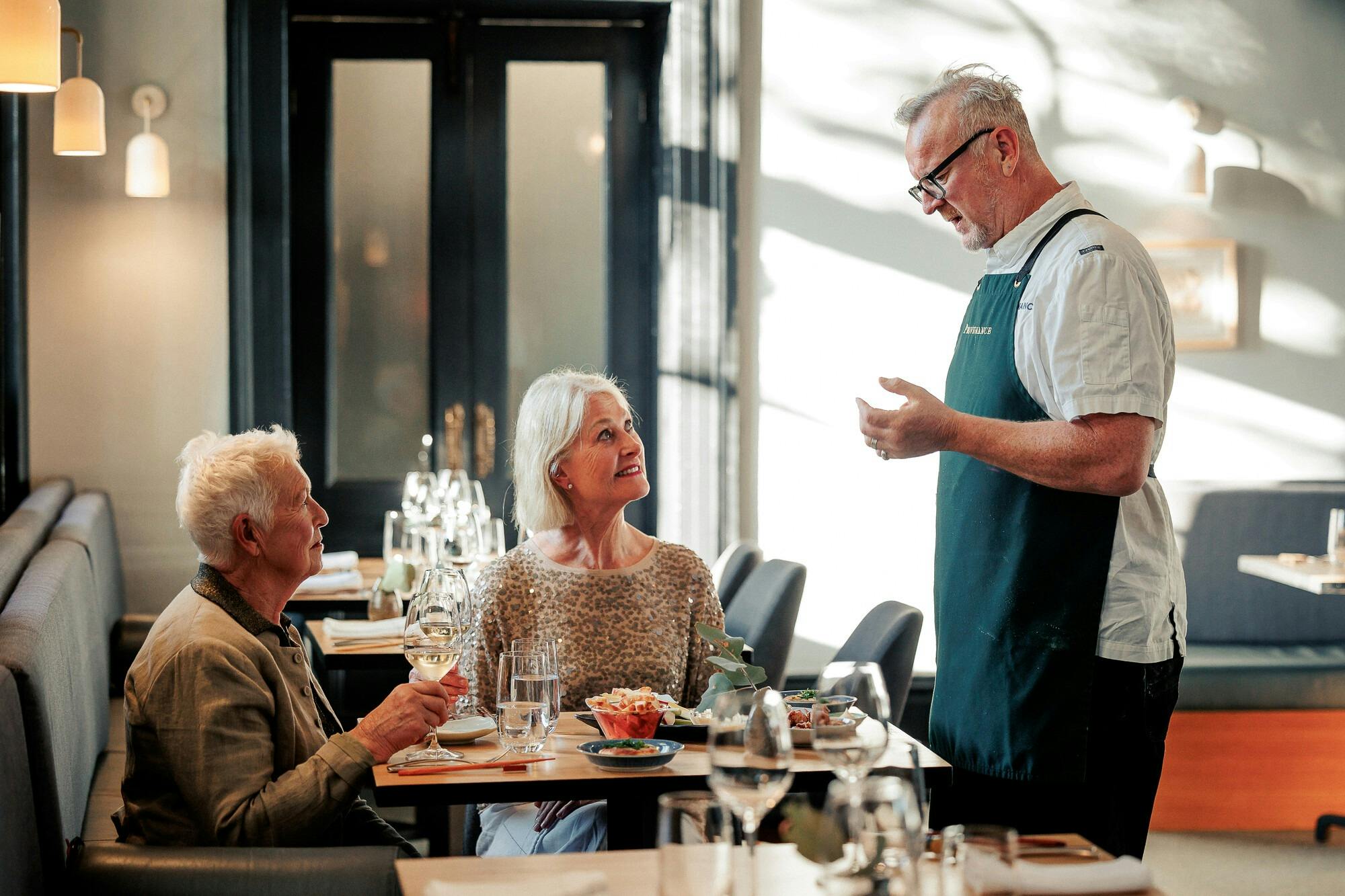 Two older women at a restaurant table speaking to the chef and drinking wine