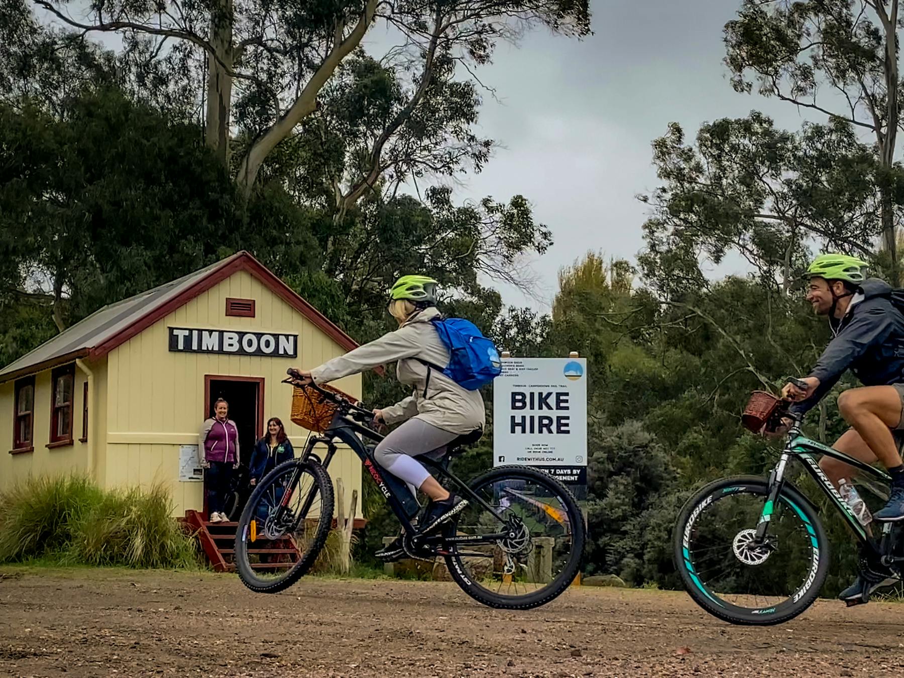 Two people riding an ebike and bicycle in front of the Timboon Railway Shed