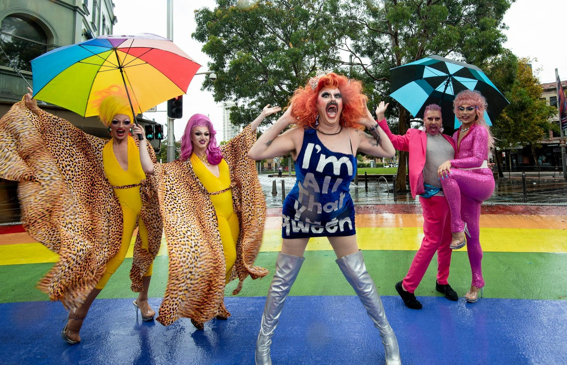 A group of drag performers in colorful, flamboyant outfits pose on a vibrant rainbow-painted street.
