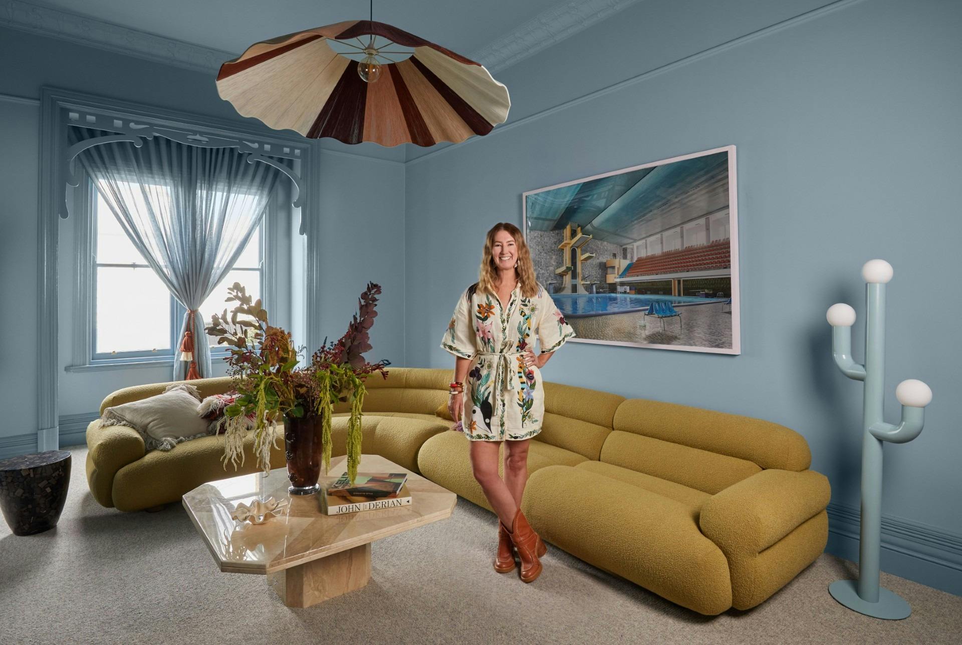 A person standing in a blue living room with a curved sofa, sculptural light and colourful wall art.