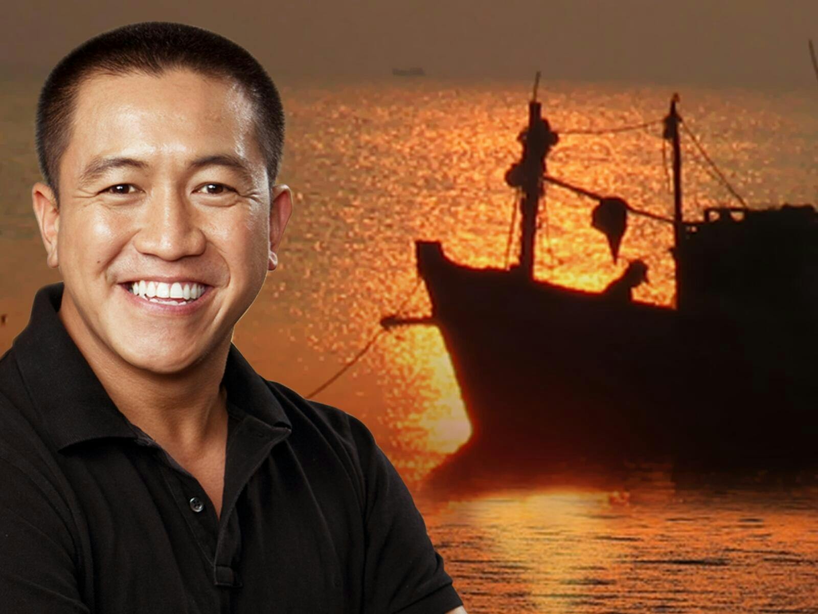 A man standing looking at the camera in front of a ship on the water