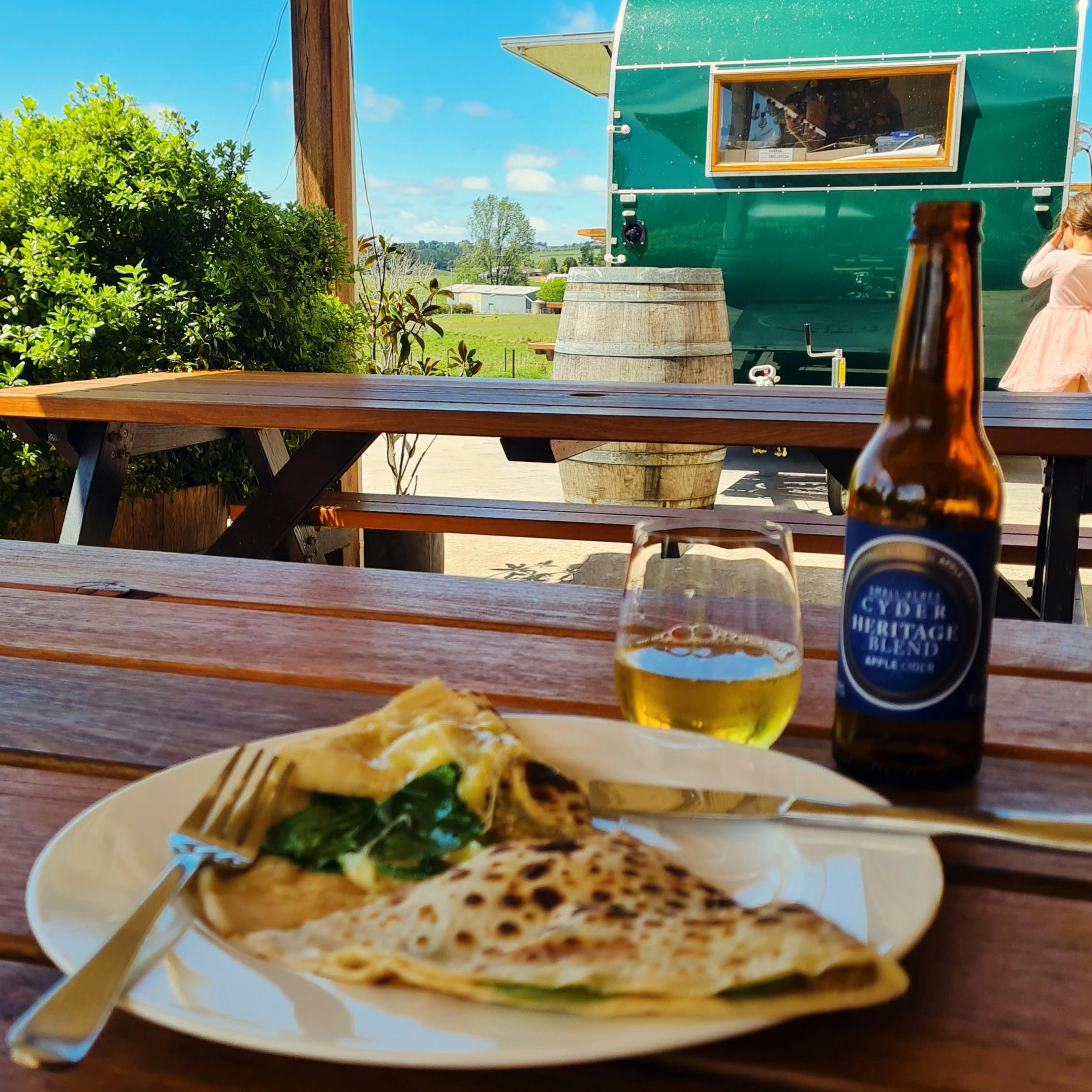 A savoury crepe and a glass of cider on a picnic table with sunny weather in the background