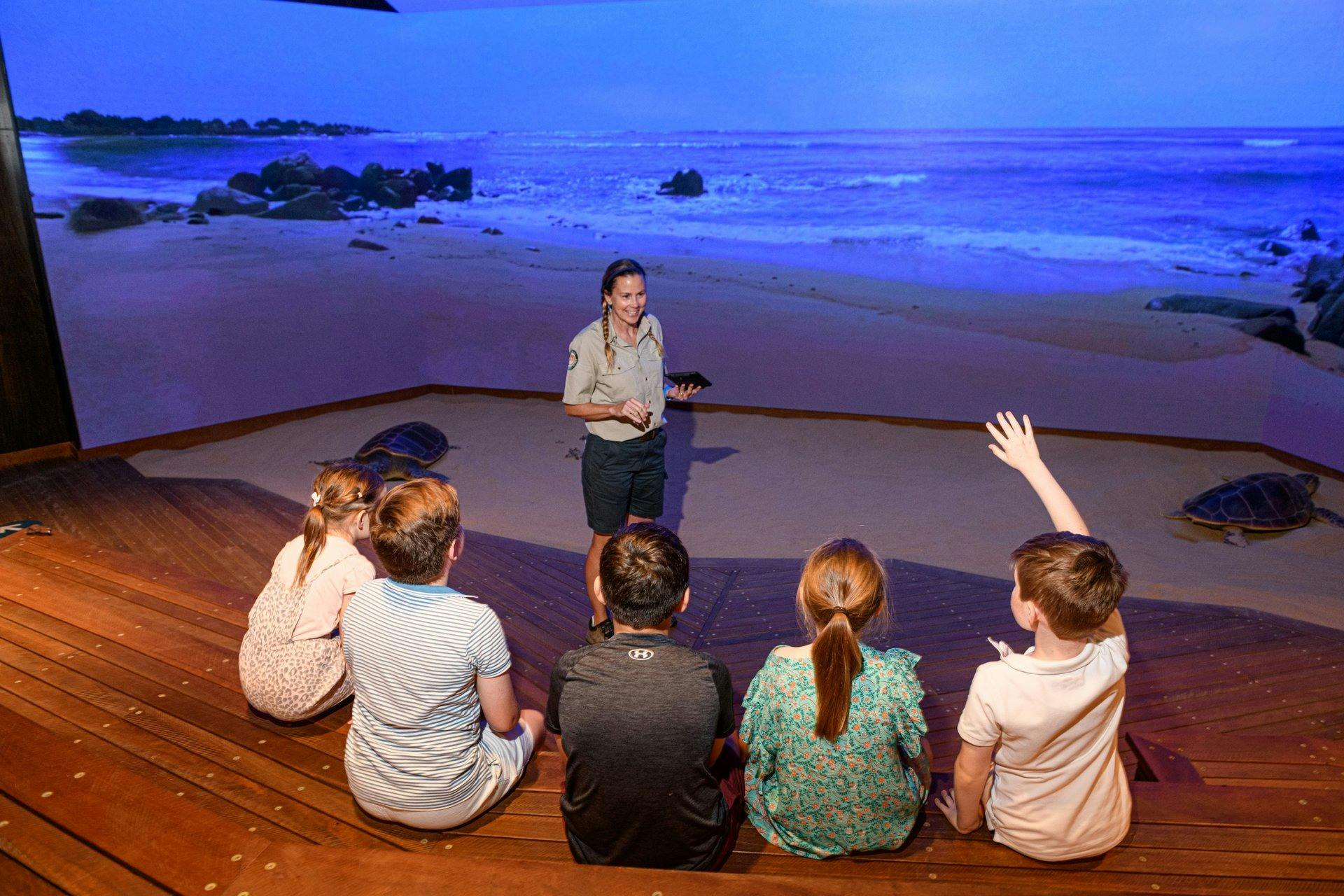 A Ranger standing in front of  a large digital screen is talking to a group of children.