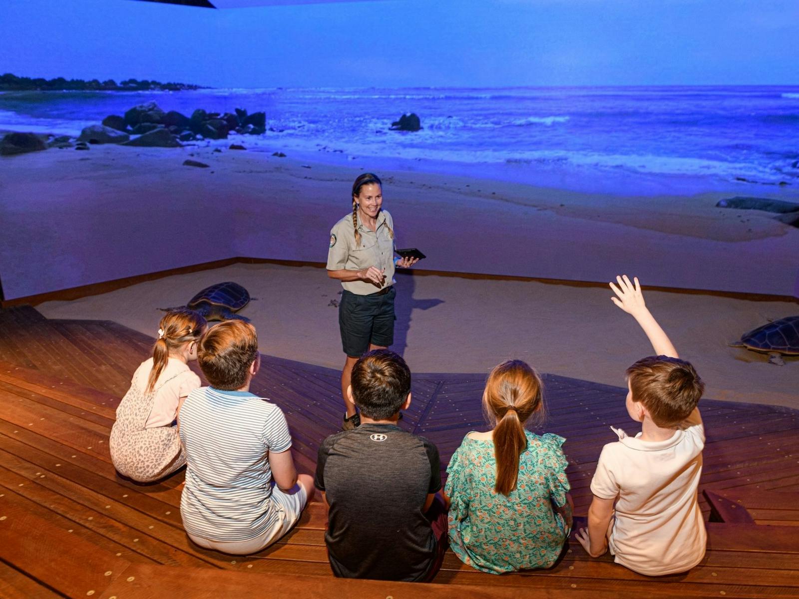 A Ranger standing in front of a large digital screen is talking to a group of children.