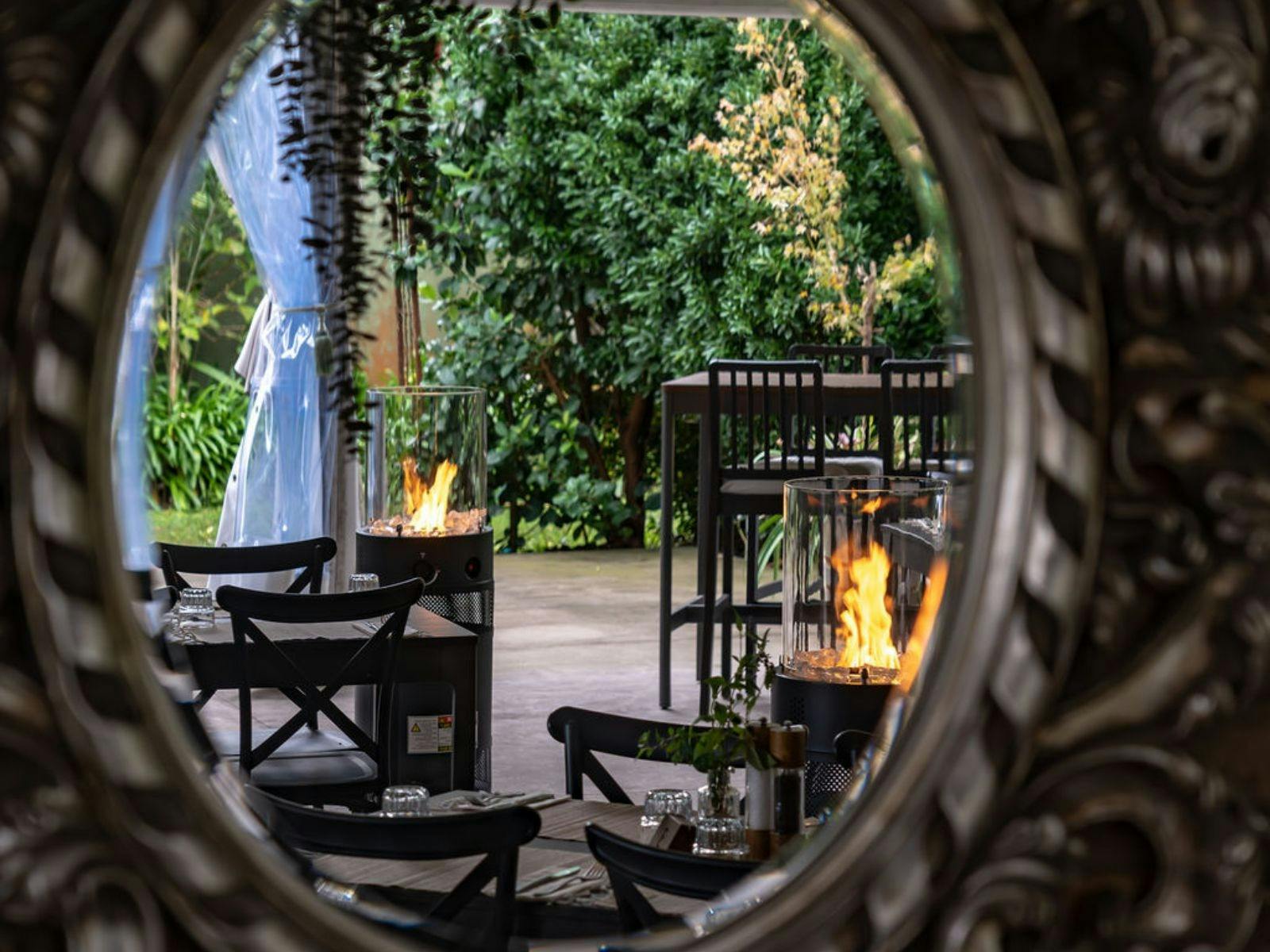 Outdoor dining area with black chairs, fire heaters, and green garden view through a round mirror.