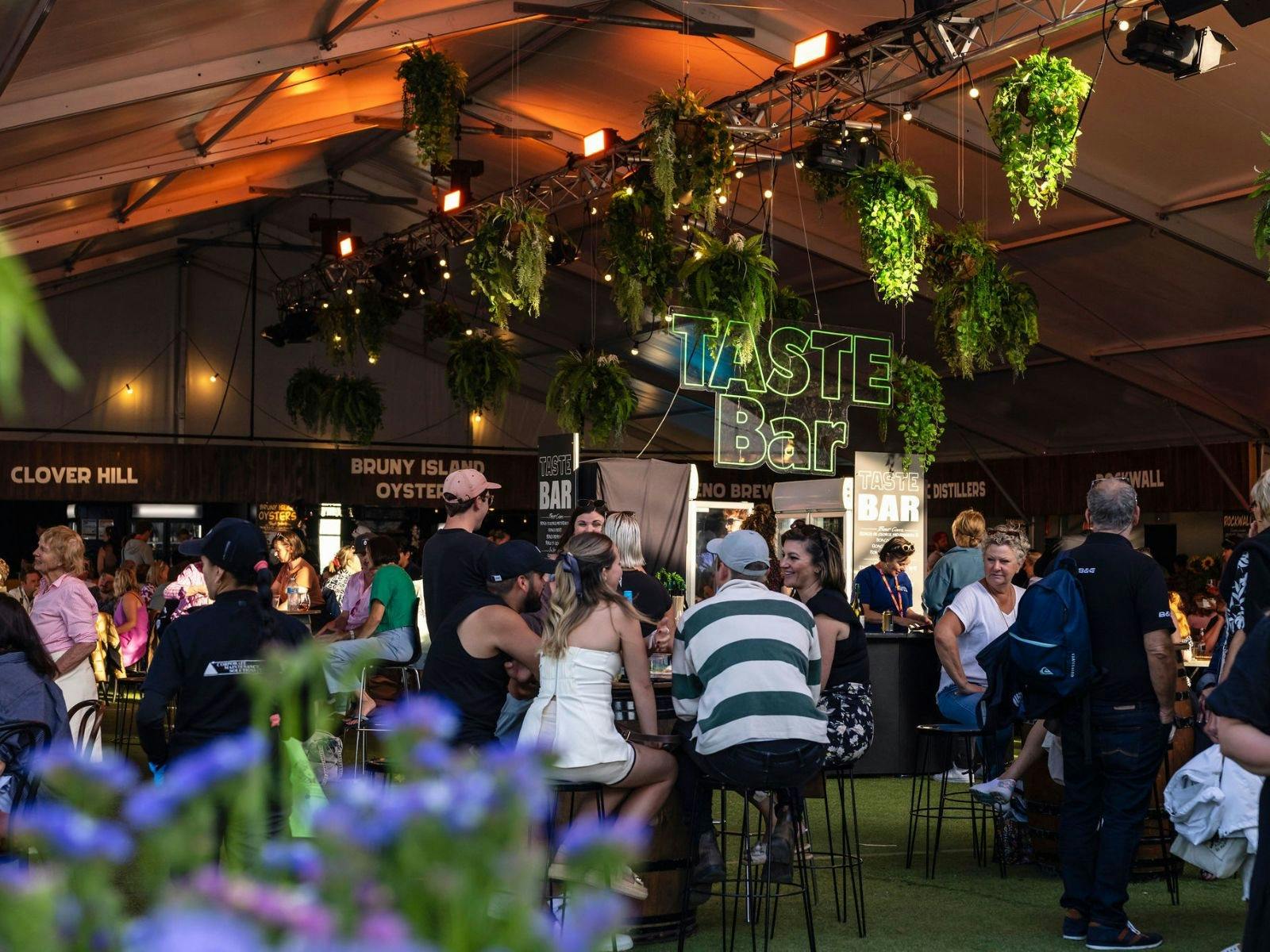 Festival-goers sit under a marquee with hanging greenery and a glowing Taste Bar sign.