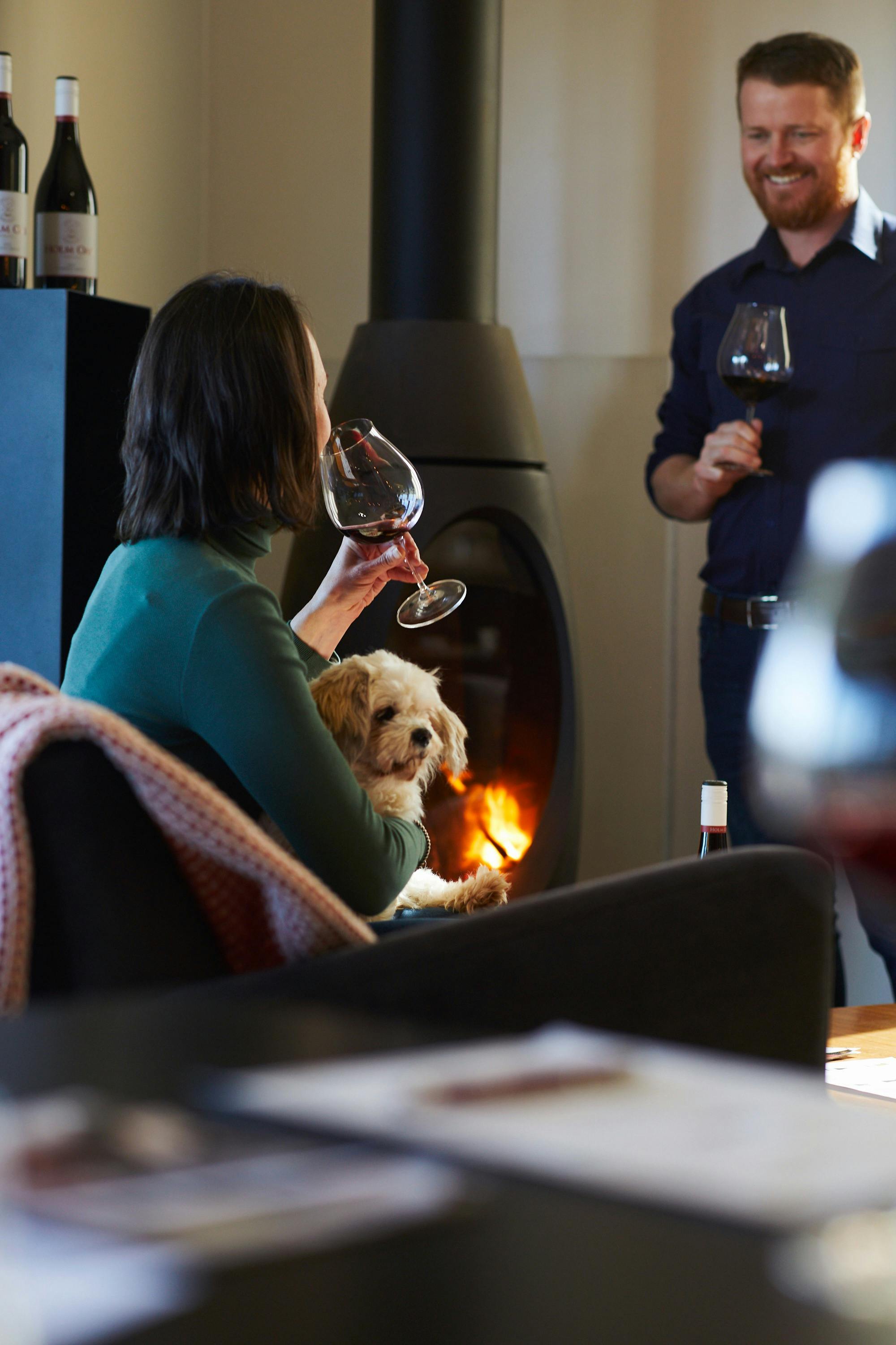 mand and woman rinking wine in front of a fire with woman holding a small white dog.