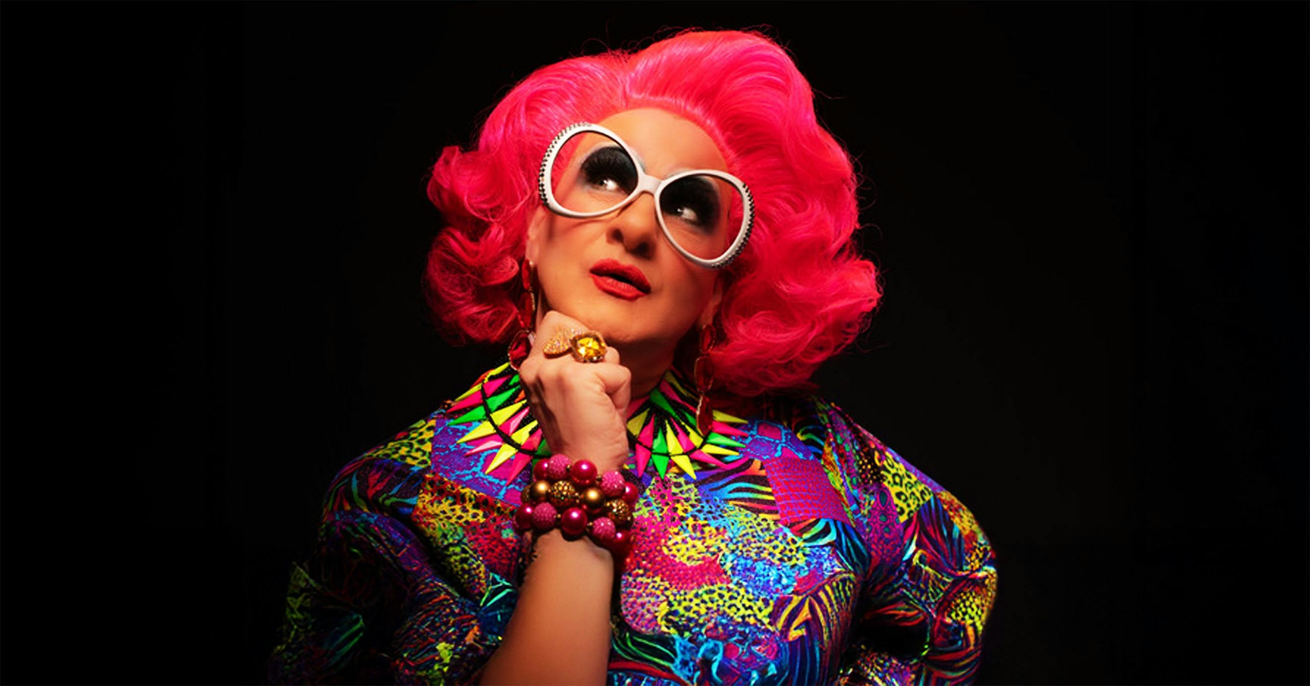 A drag queen wearing quoffed shoulder length wig, big glasses, and bright colours, in thought.