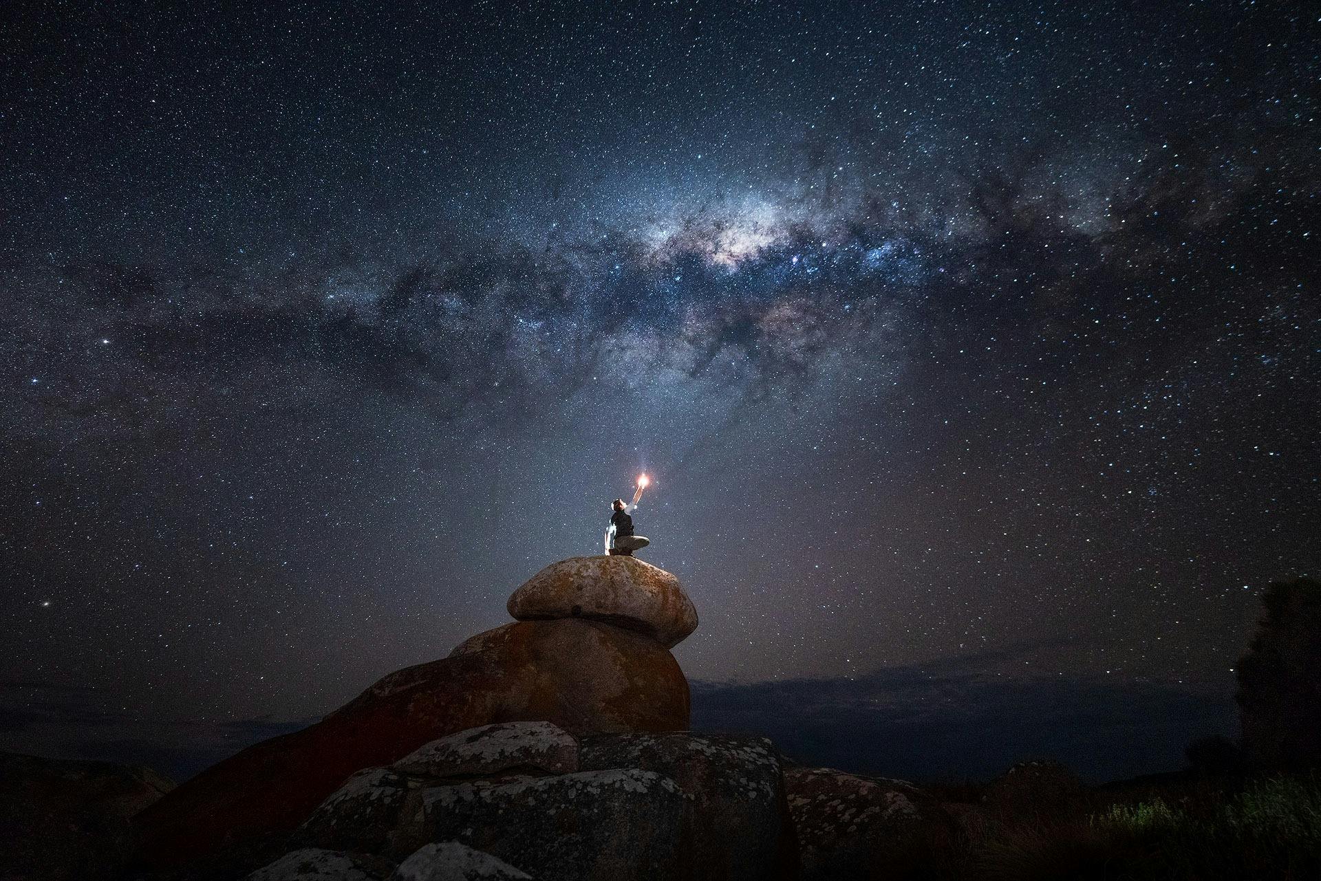 A person sits atop a rock formation at night hold a torch light towards the milky way.