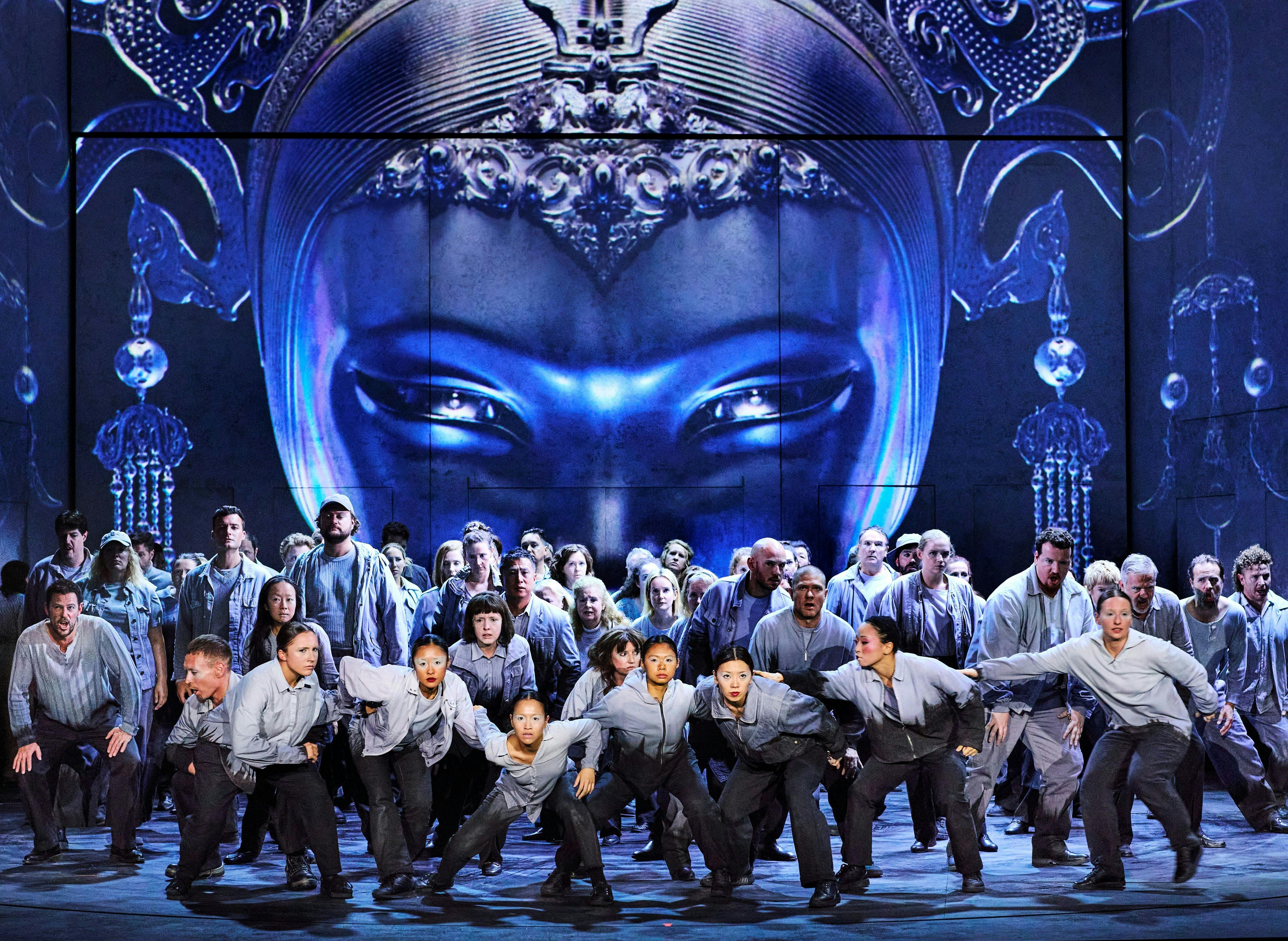 Large ensemble in gray costumes onstage before a giant blue Chinese avatar face.