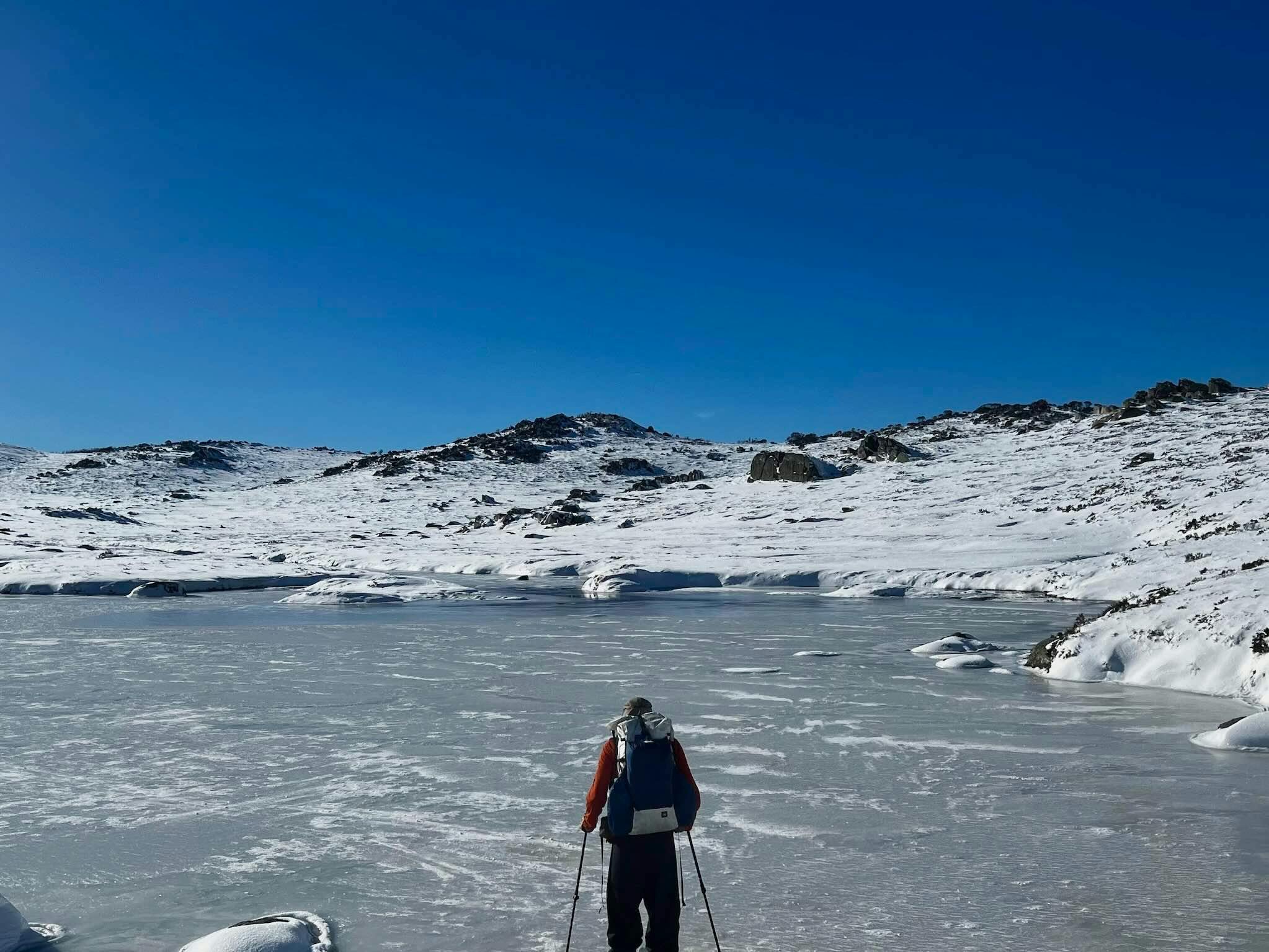 Ein Wanderer überquert einen zugefrorenen See, in der Ferne blauer Himmel.