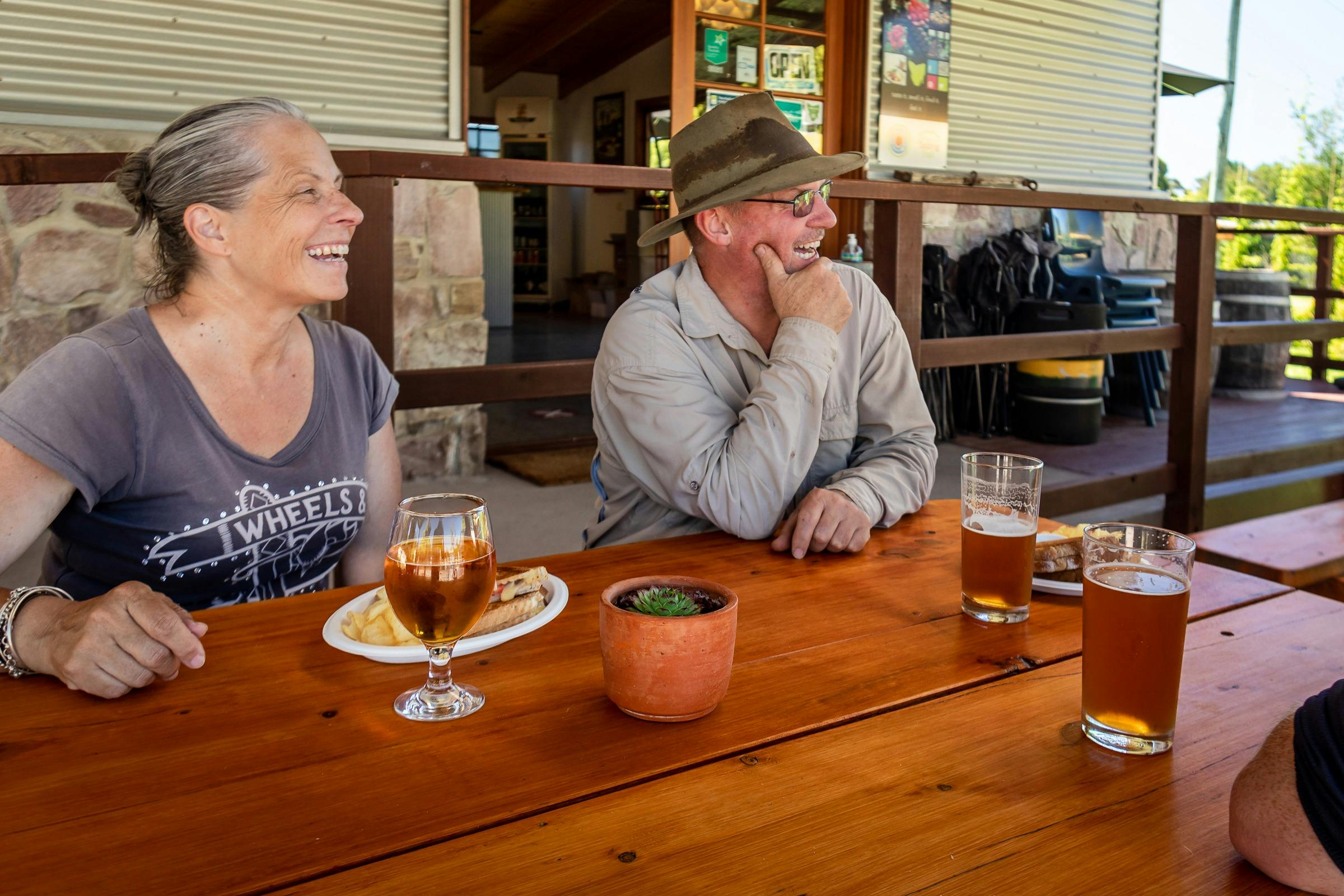 Woman and man with toasted sandwiches and beers sitting at a picnic table in the shade.