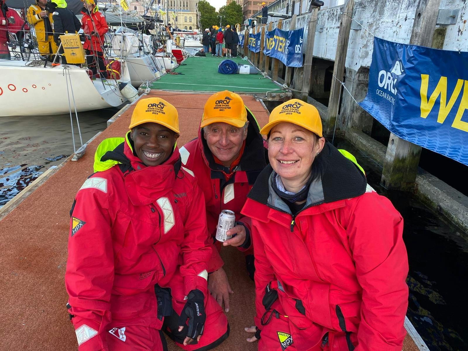 Three smiling Melbourne to Hobart sailors in wet weather gear kneeling on the pier in Hobart
