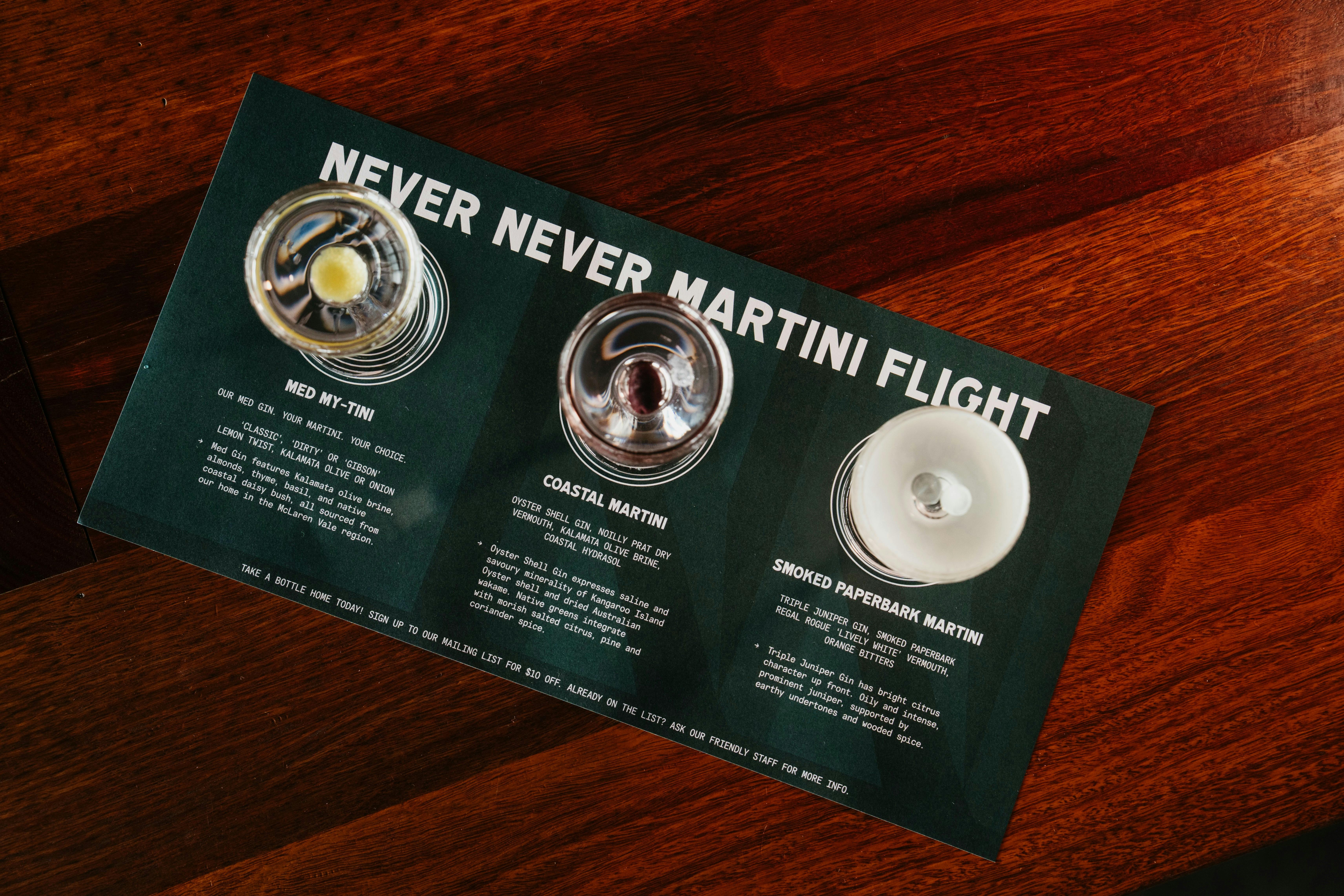 Three miniature martinis are shown on a dark green tasting flight mat with white writing.