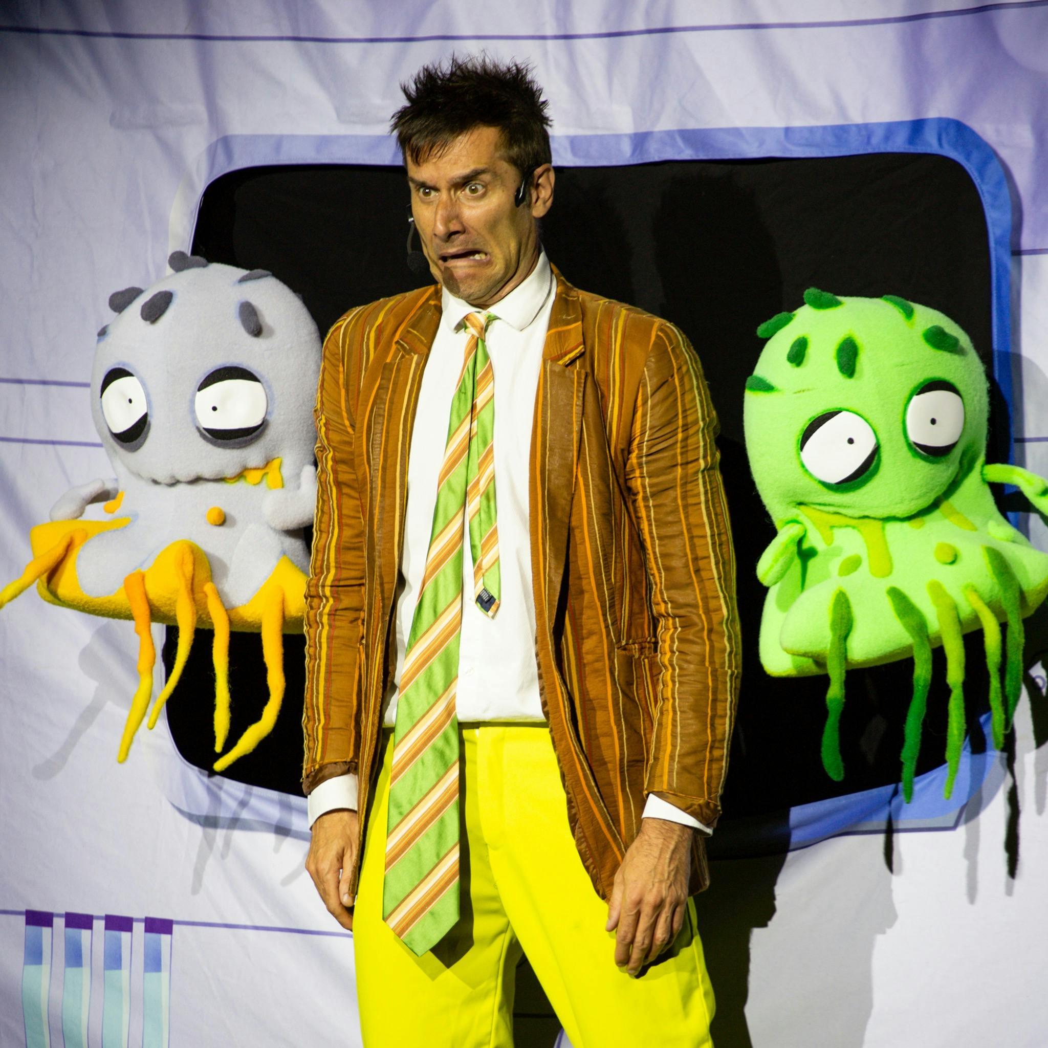 Man in bright colours looking scared next to two snotty zombie puppets