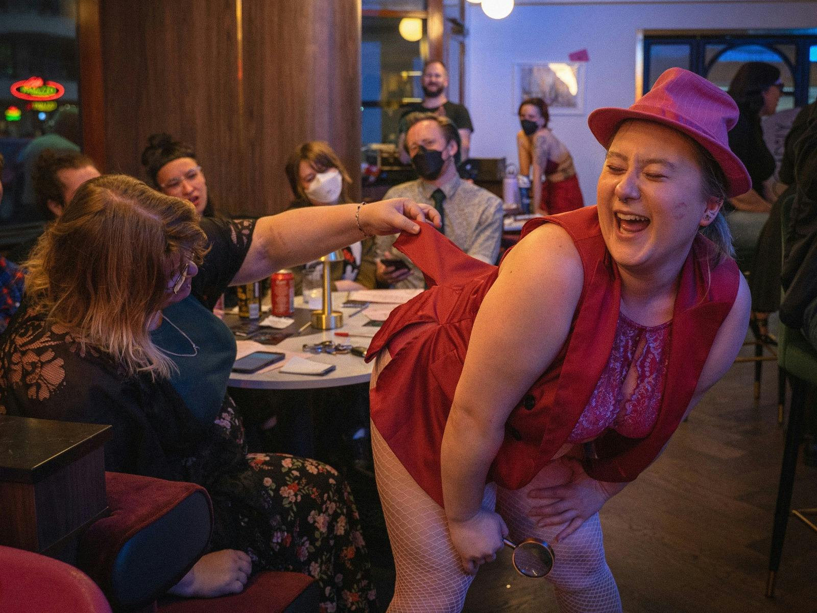 an NB performer in pink bends over while a woman lifts their costume