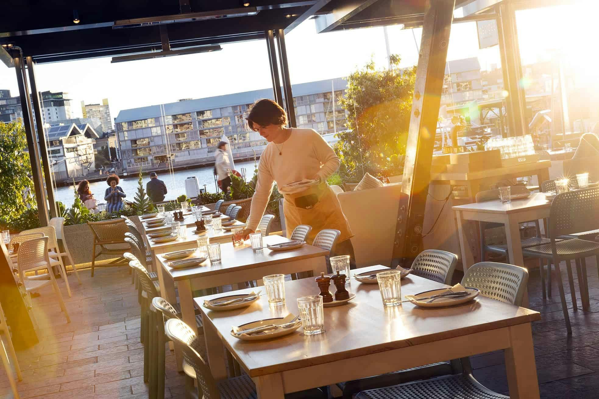 A staff member sets tables on the waterfront terrace with the water and wharf buildings behind.