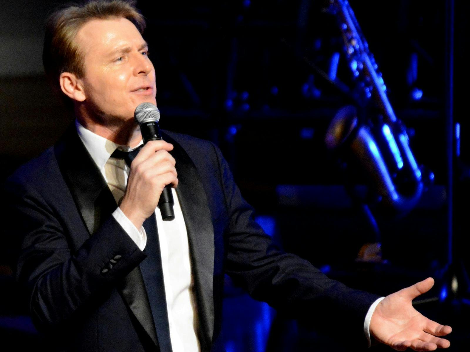 Well-dressed gentleman singing into a hand-held microphone,  with a blue lit trombone in background