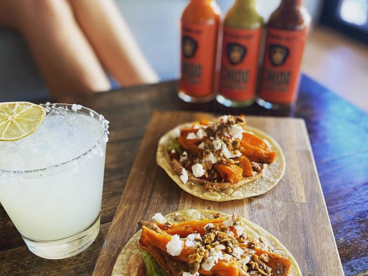 a margarita and tacos on the wooden board with 3 sauces in the background