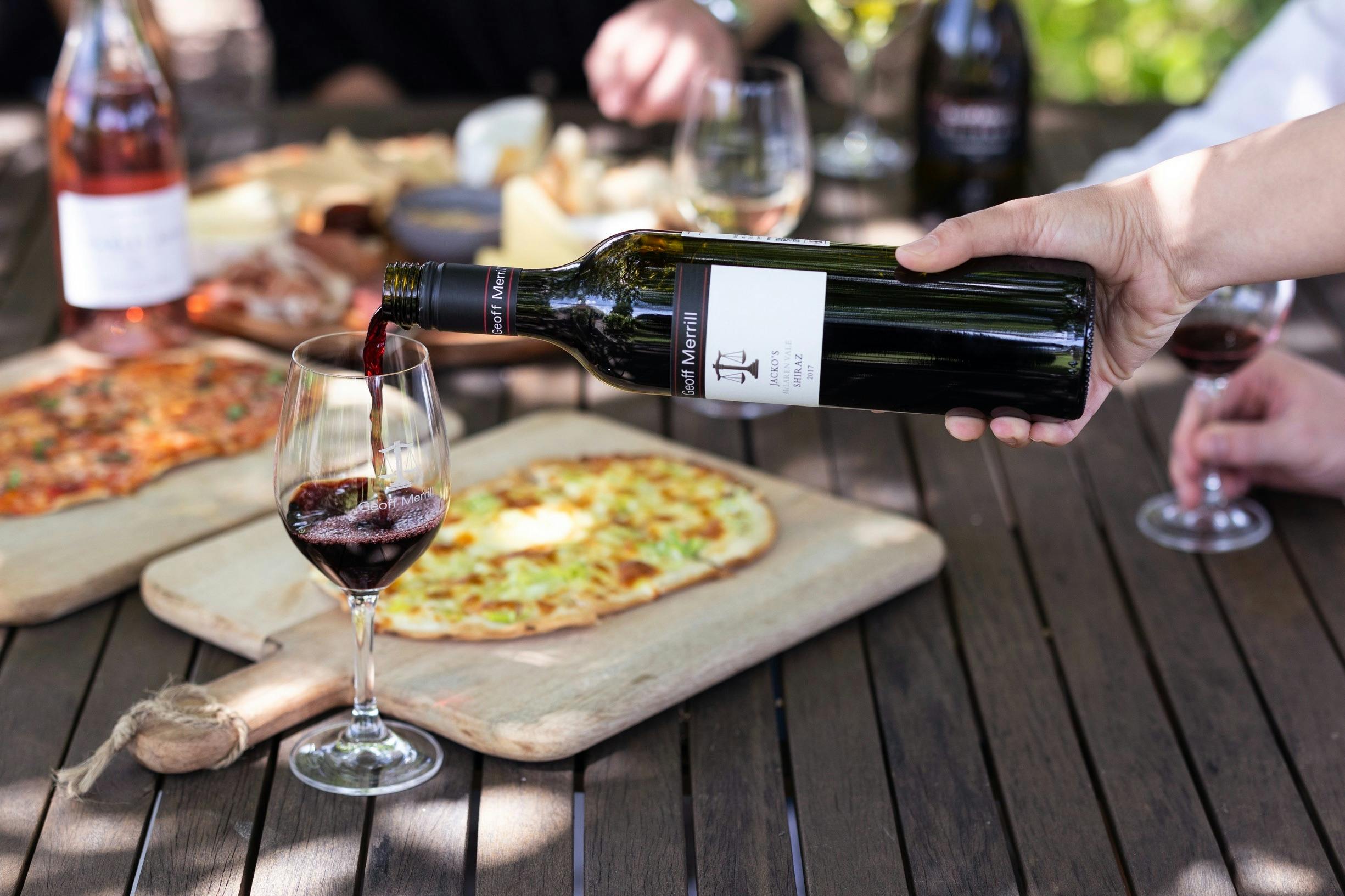 A glass of red wine being poured with a flammekueche behind
