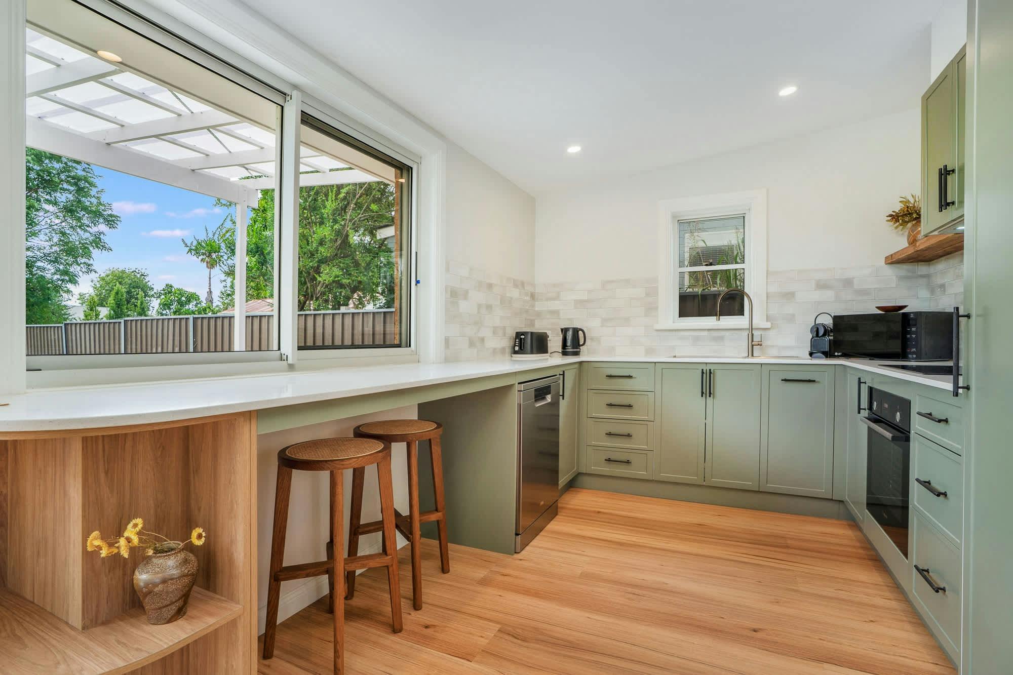 Kitchen with green cabinets, wooden shelfs, white bench tops and wooden stools