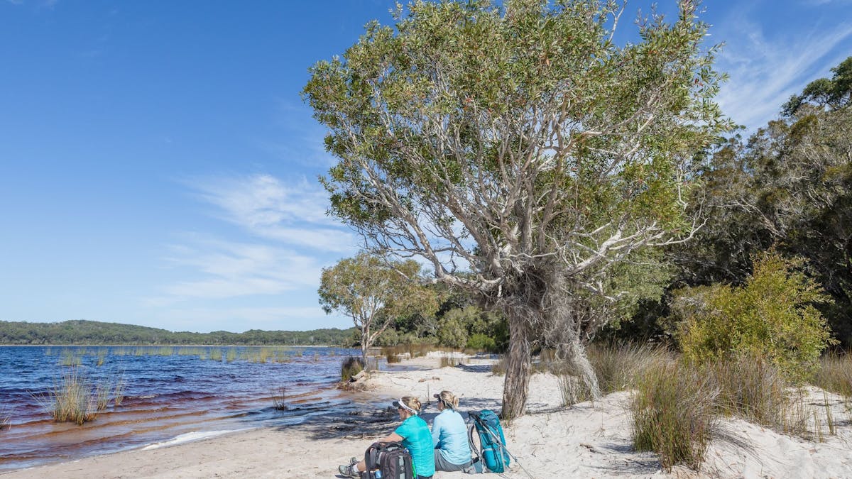 photo showing two hikers sitting on a sandy beach at Lake Birrabeen