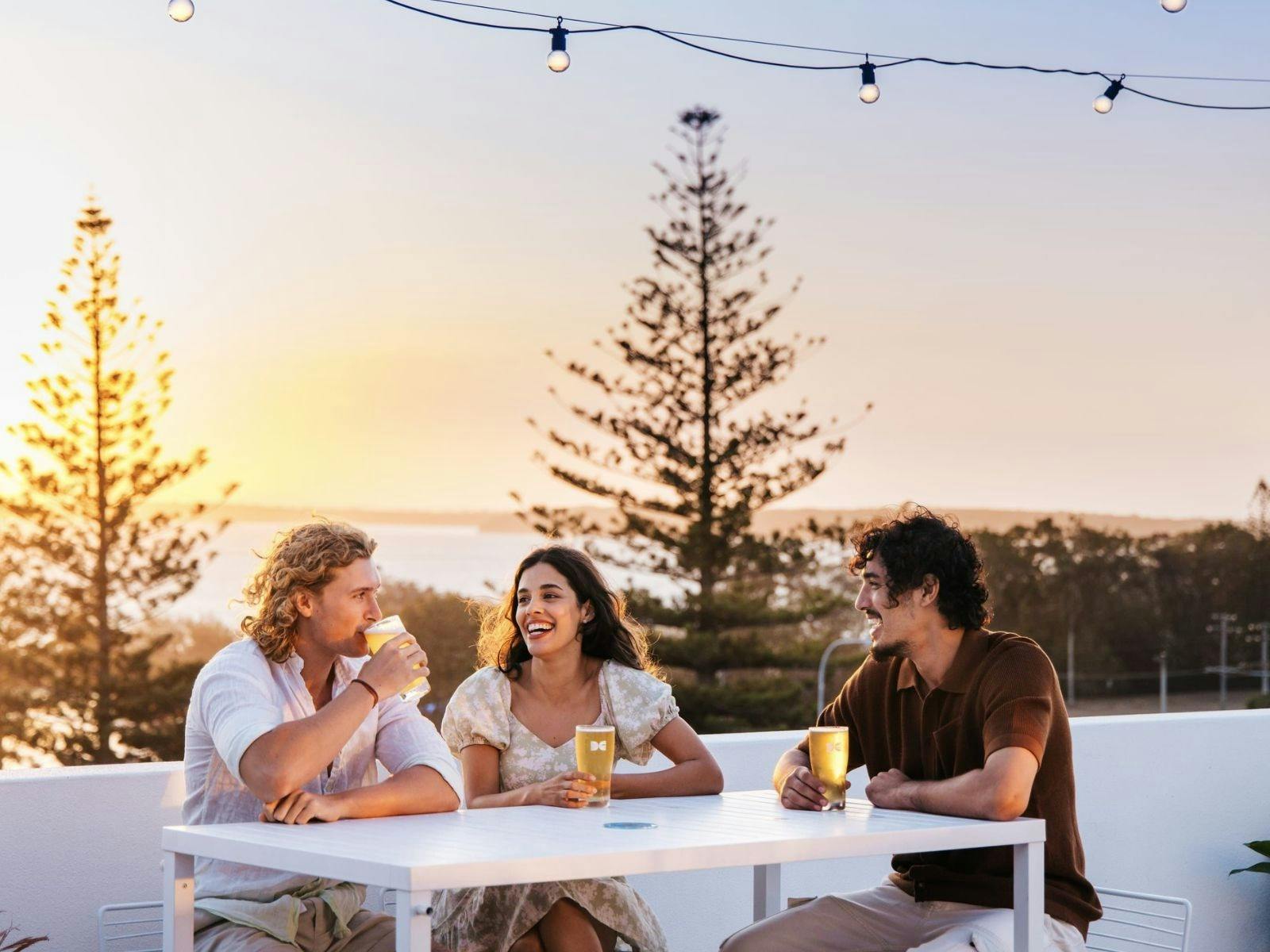 Three people drink beers at a white table, background is silhouette of trees