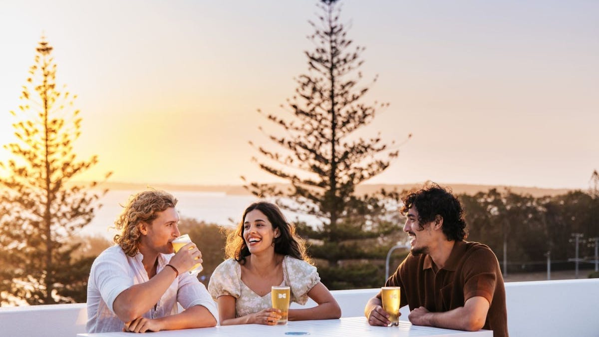 Three people drink beers at a white table, background is silhouette of trees