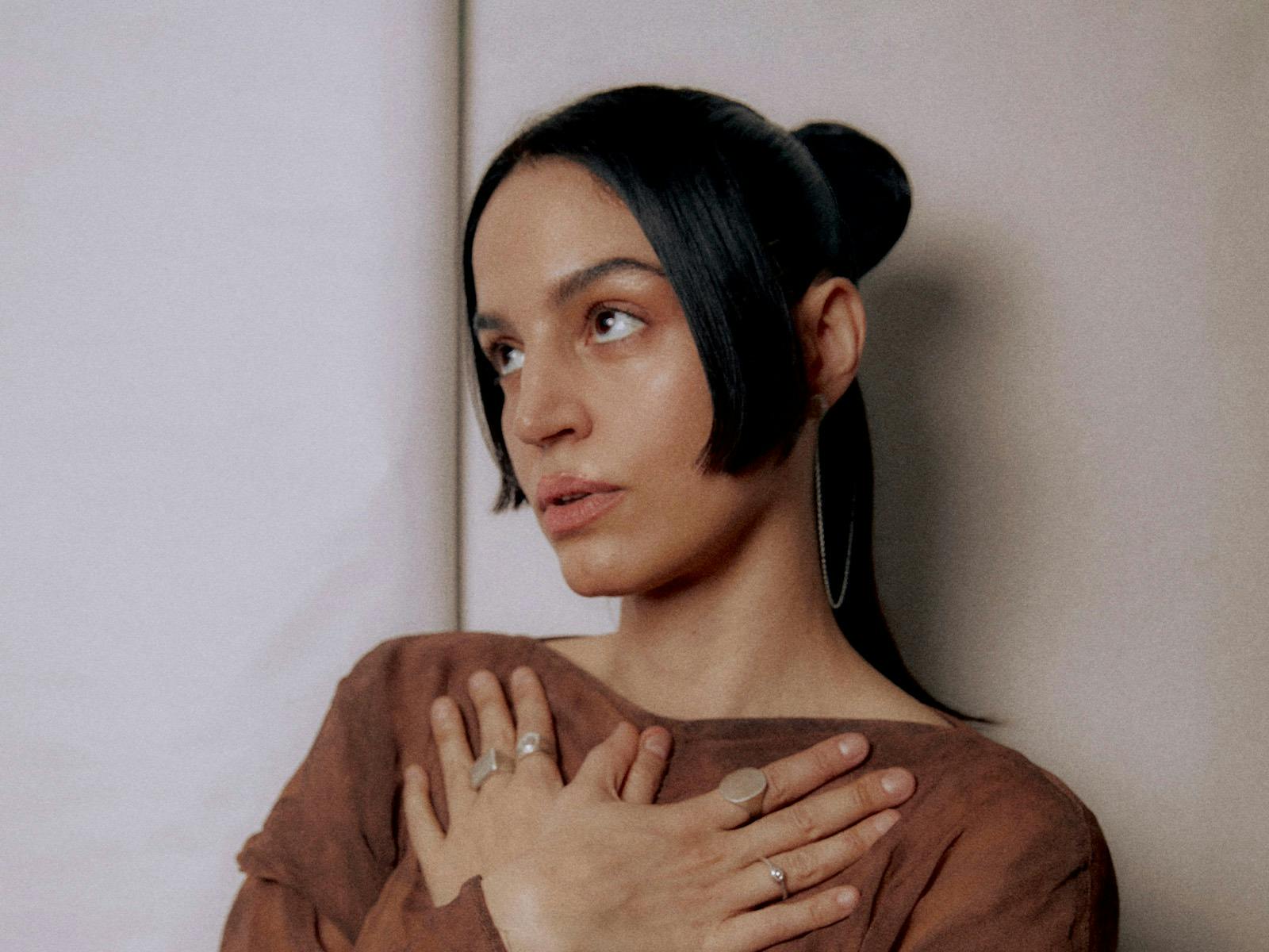 Maria Arnal standing by cream background holding hands to heart