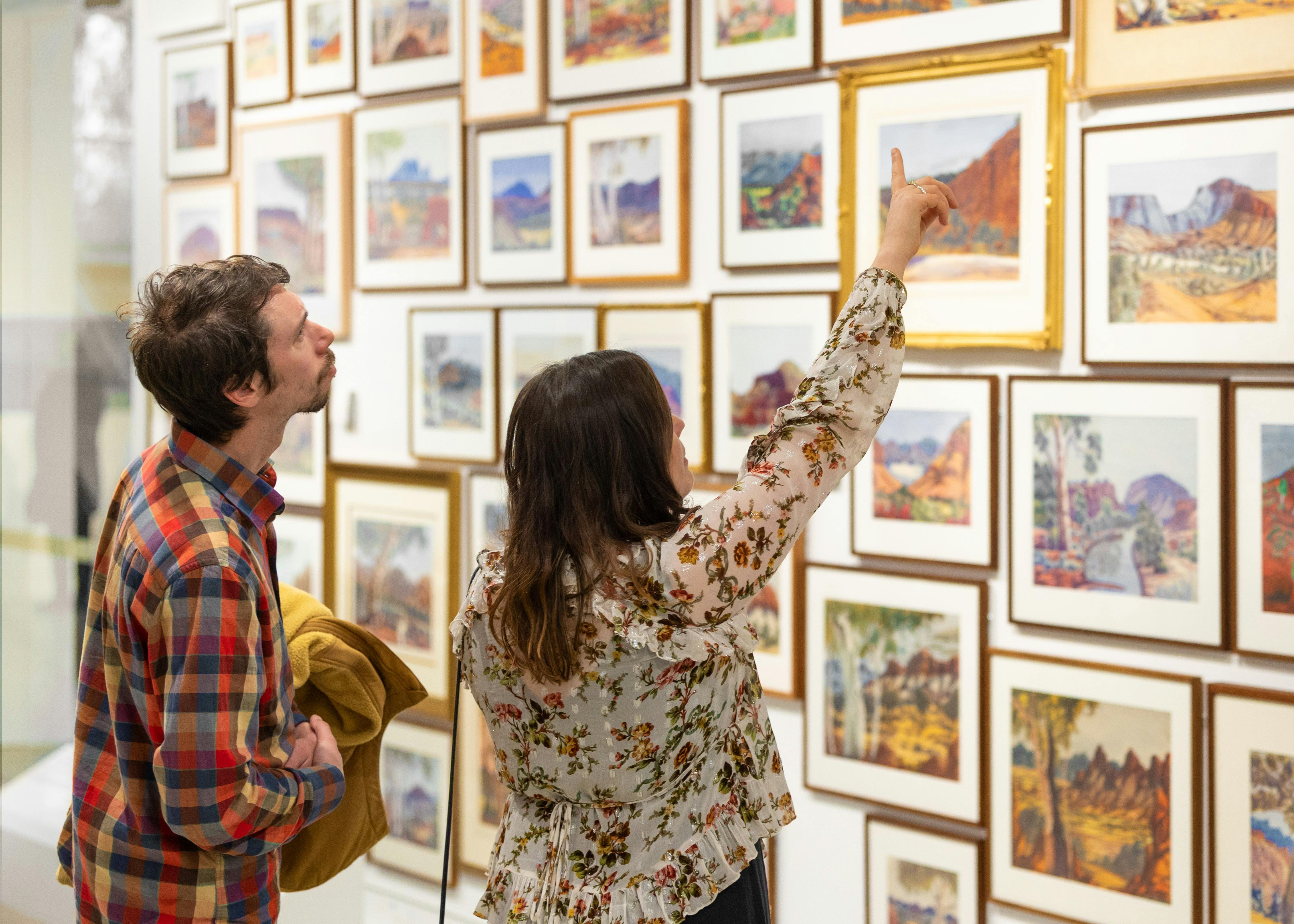A man and woman point at artworks hung salon-style in a gallery.