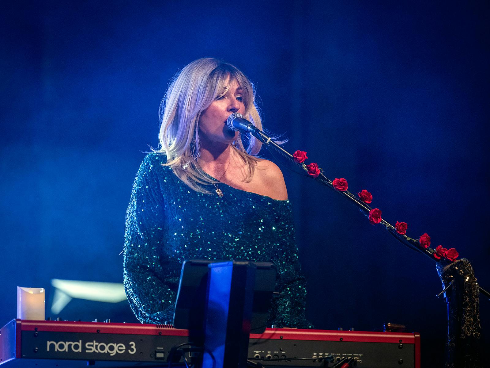 Christine McVie impersonator singing and playing keyboard on stage witha dark blue smokey background