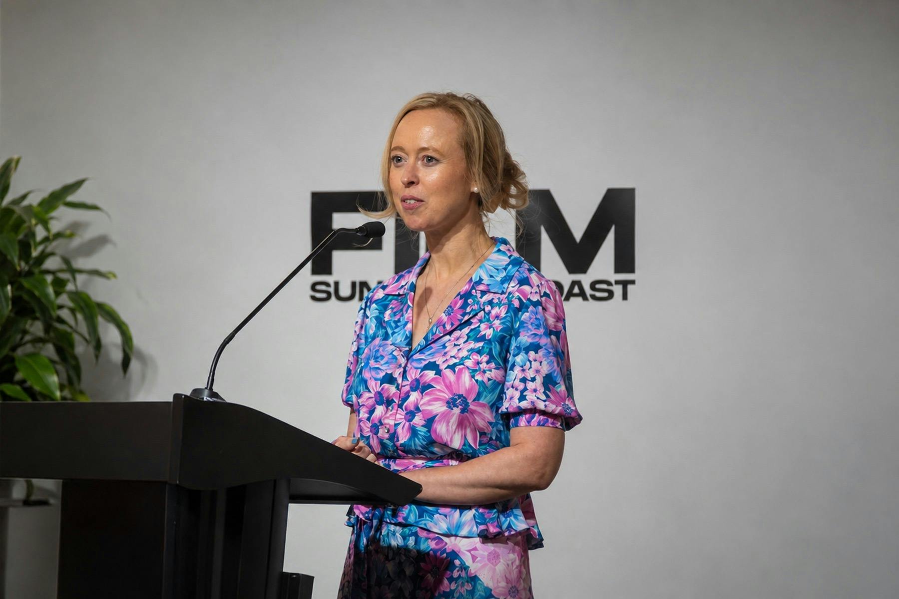 Picture of woman in floral dress speaking at podium with FILM Sunshine Coast Behind her
