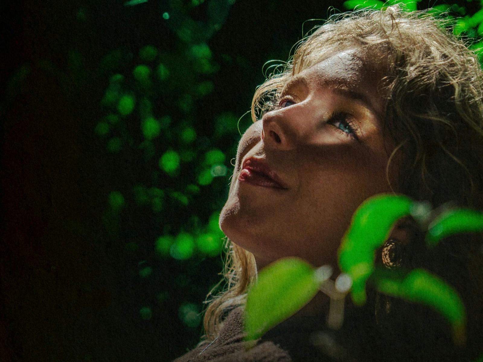 A close up of a blonde woman standing amongst greenery with her face turned to the light