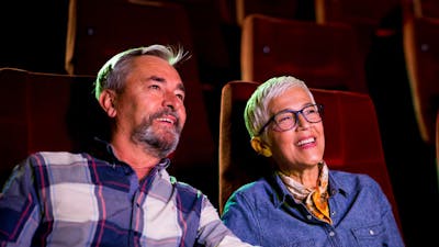 A man sitting on left and woman on right, both sitting at the cinema looking past the camera